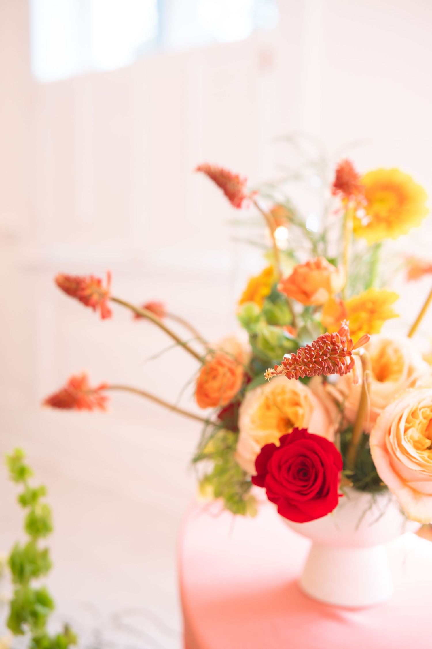 Vibrant orange, yellow, and red rose arrangement in white vase on a pink table.