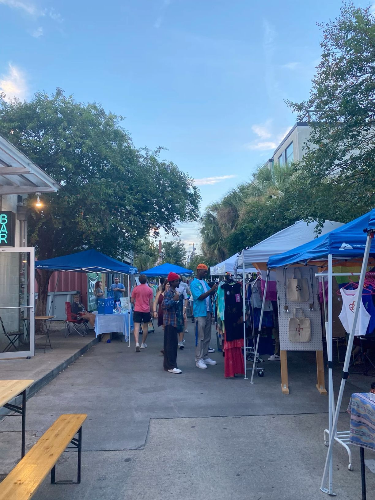 Outdoor market with vendor tents, shoppers browsing goods, and lush trees under a bright blue sky.