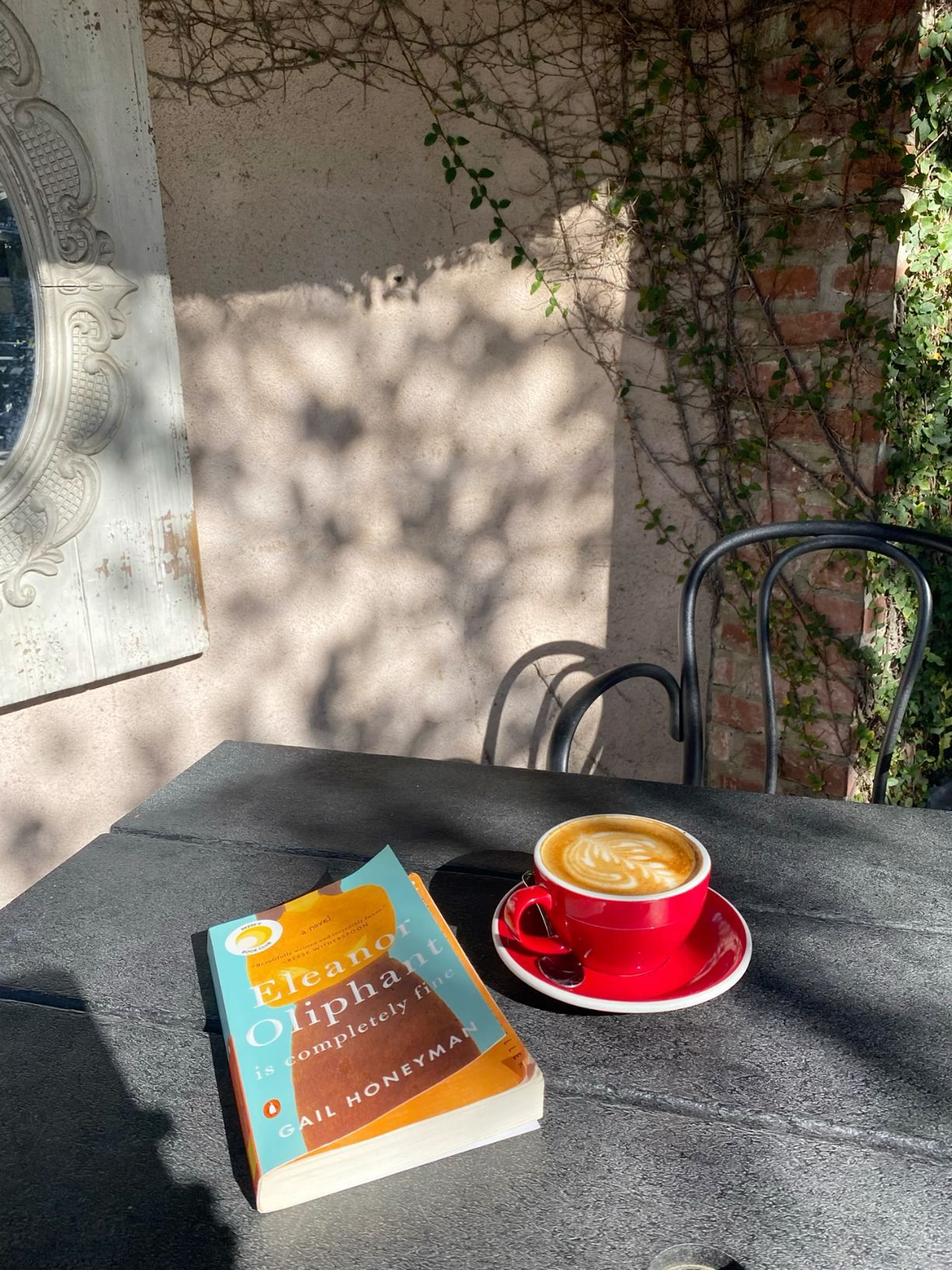 Latte in red cup beside Eleanor Oliphant novel on outdoor cafe table with ivy wall.