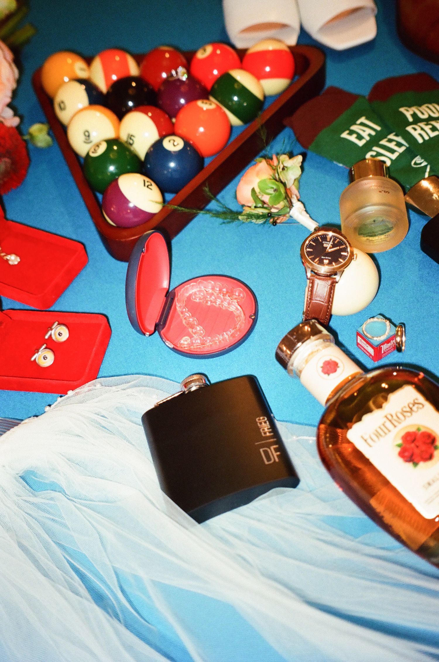 Flat lay of billiard balls, flask, Four Roses bourbon, watch, and jewelry on a blue surface.