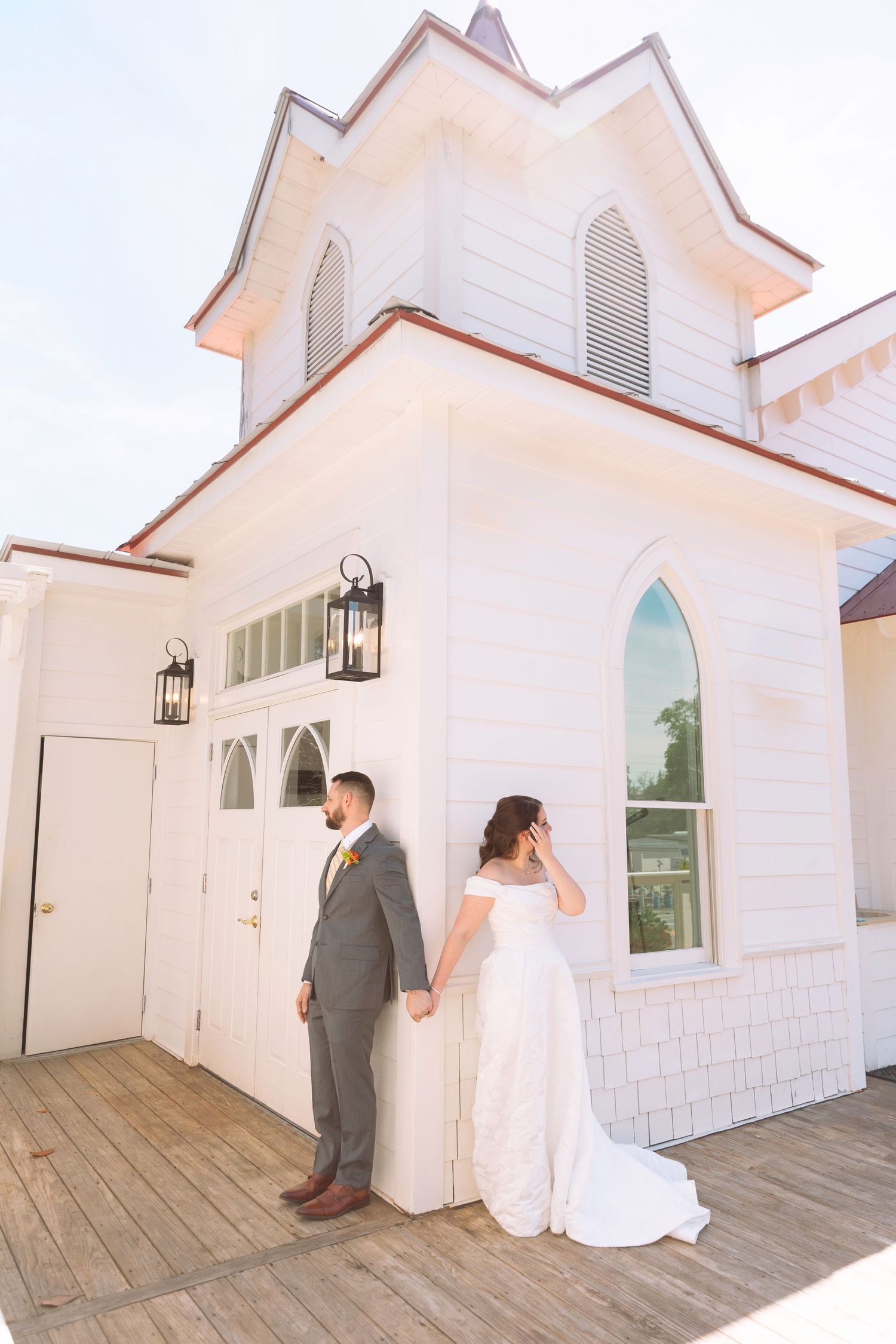 Bride and groom standing apart against a white chapel exterior on a wooden deck, bride in off-shoulder gown, groom in gray suit.