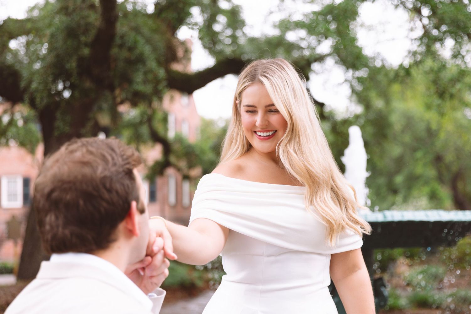 Woman in white off-shoulder dress smiling during outdoor marriage proposal near a fountain.