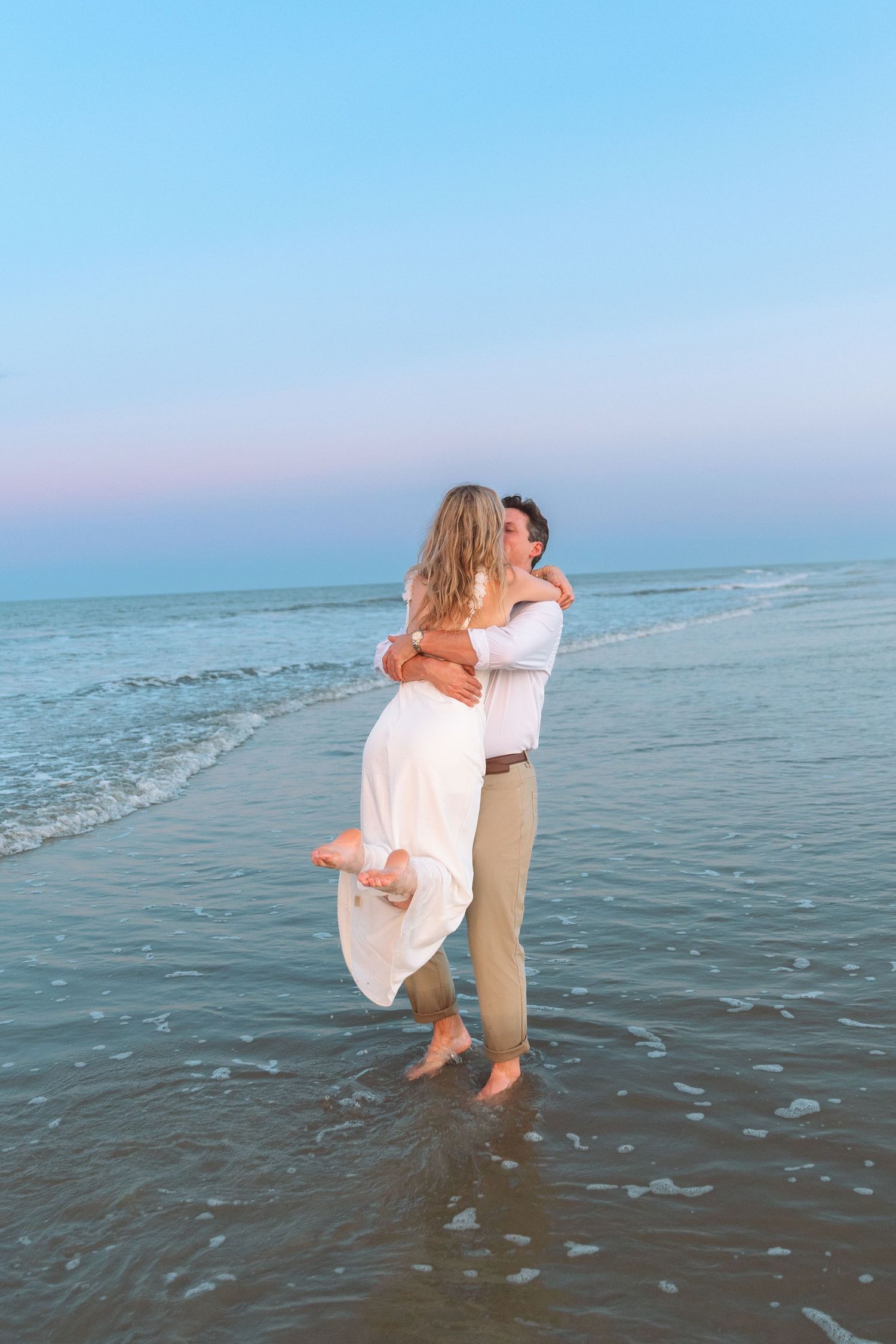 Man lifting woman in white dress on beach at twilight, romantic couple embracing in ocean waves.