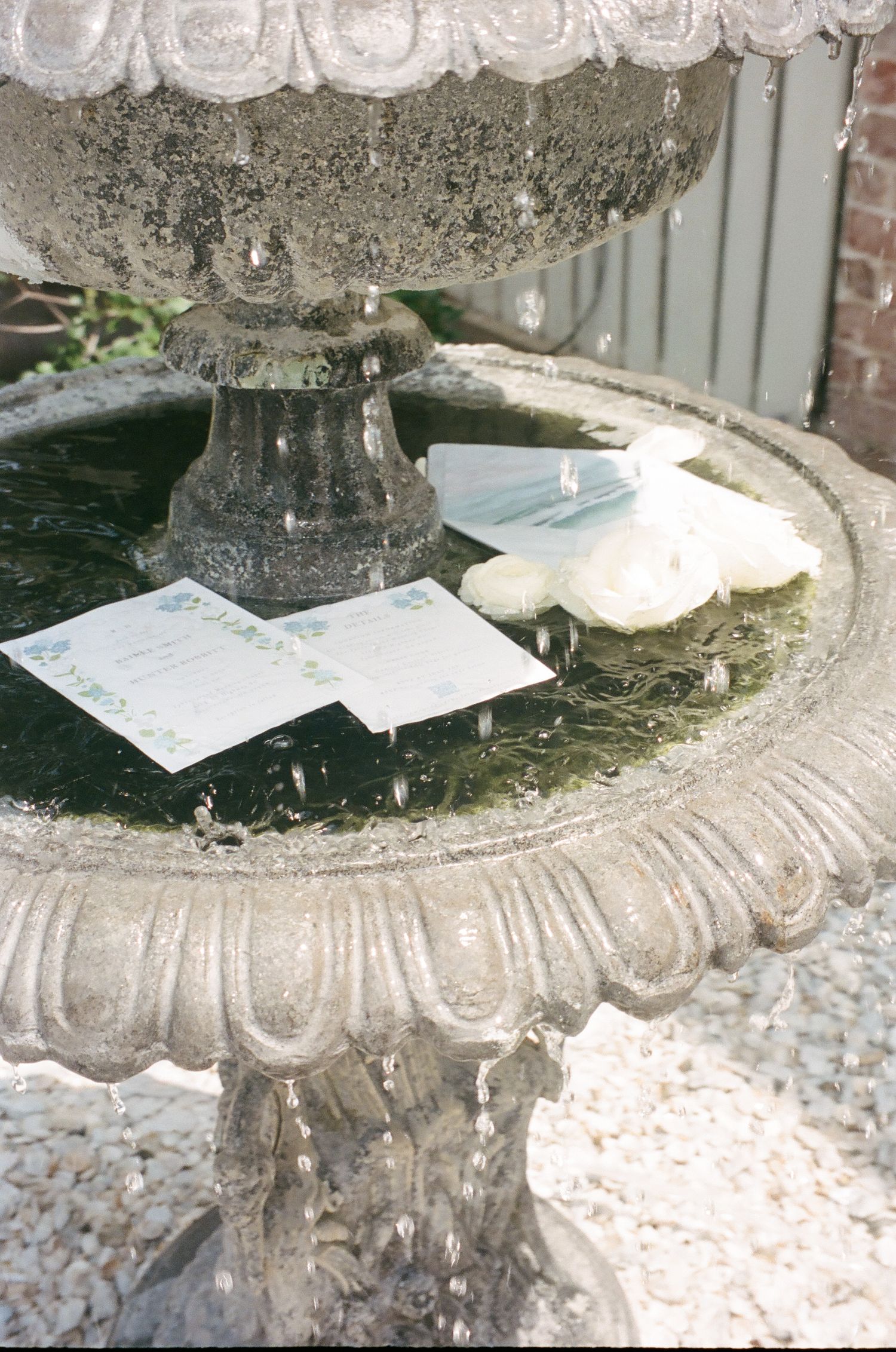 Stone tiered garden fountain with wedding invitations and white roses resting on its basin outdoors.