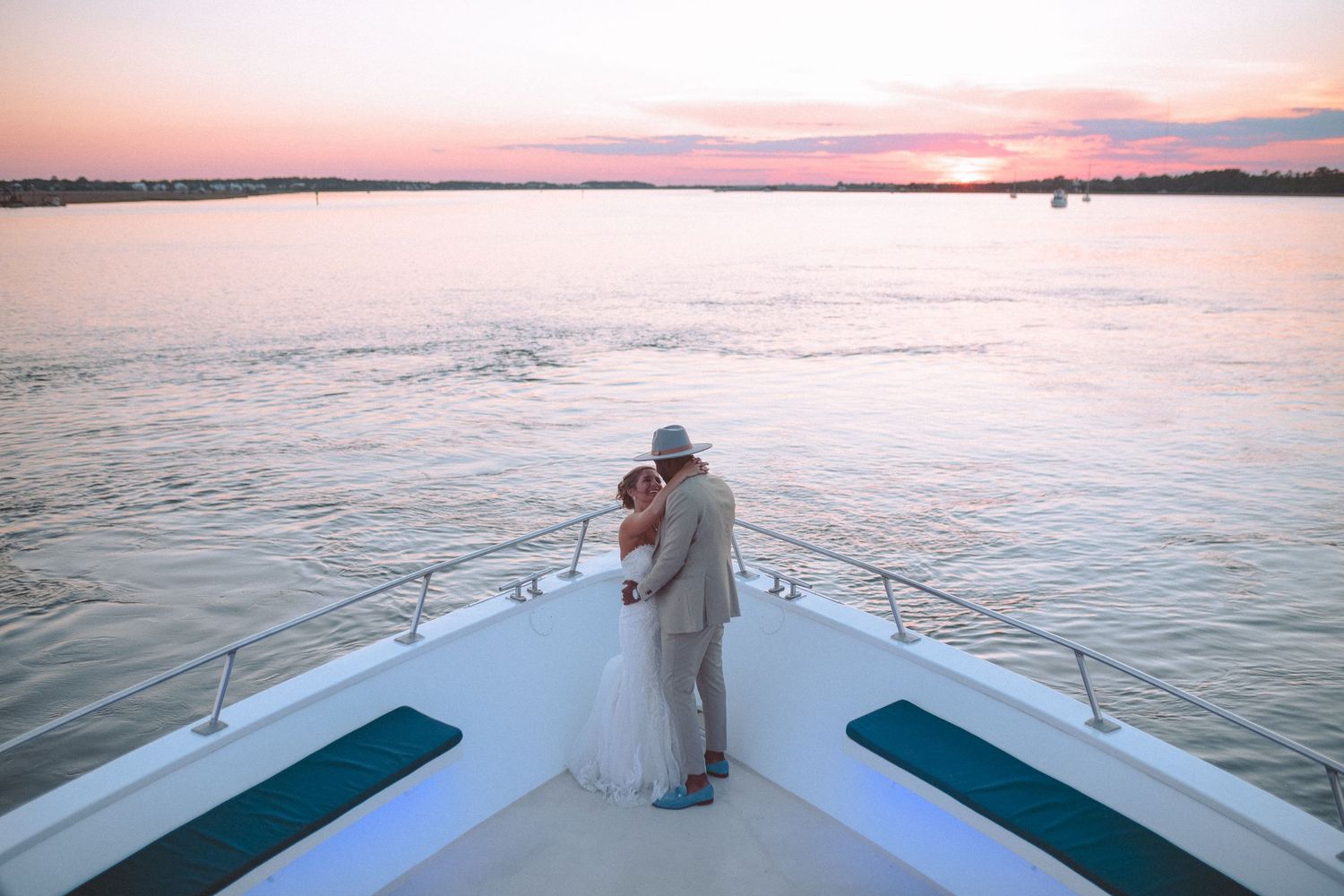Newlywed couple embracing on the bow of a white yacht during a beautiful pink and orange sunset.
