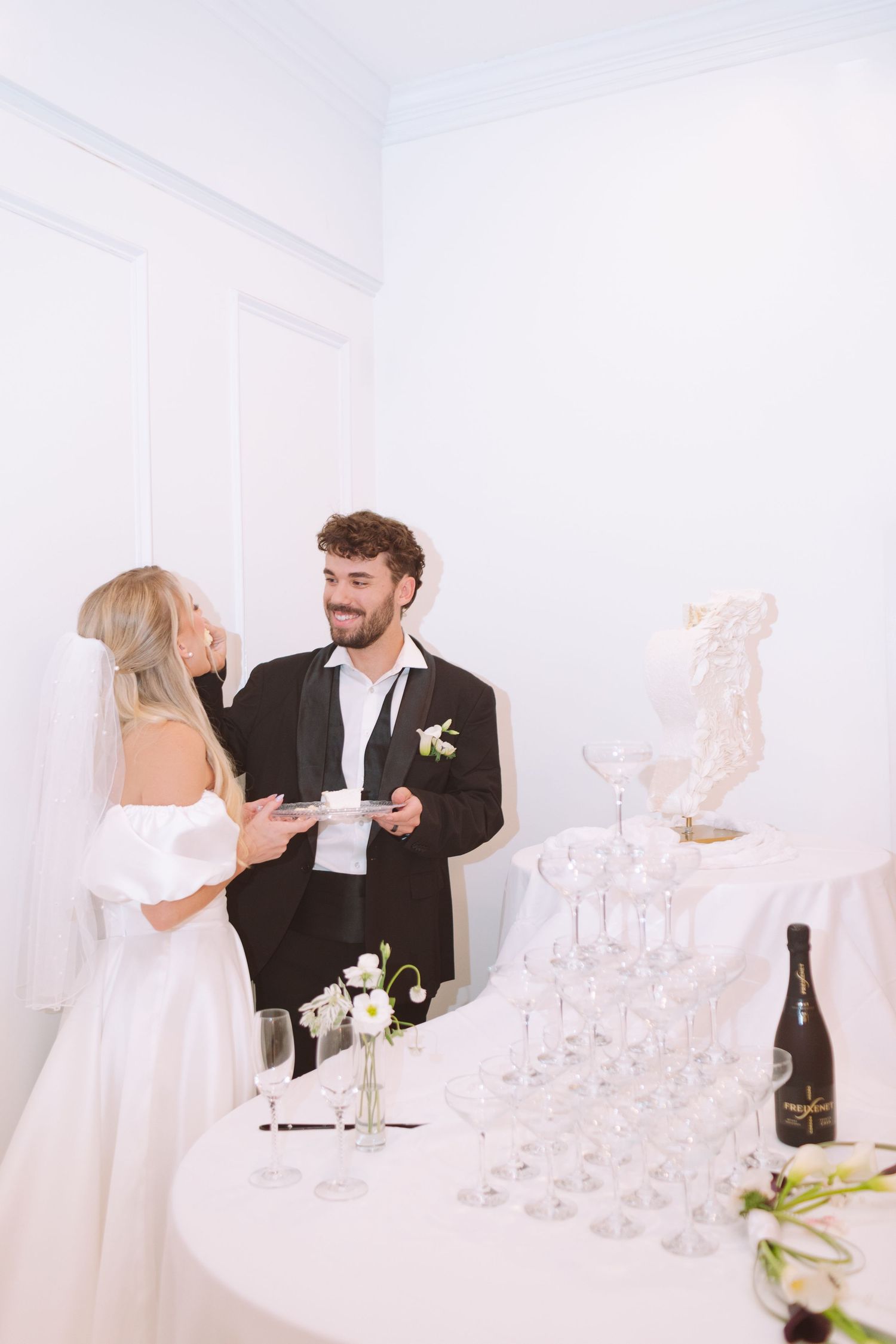 Bride and groom sharing wedding cake slices beside a champagne tower at their white-themed reception.