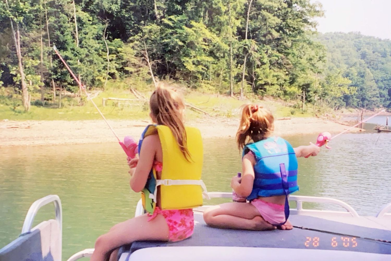 Two girls in life jackets fish from a boat on a calm lake surrounded by trees.