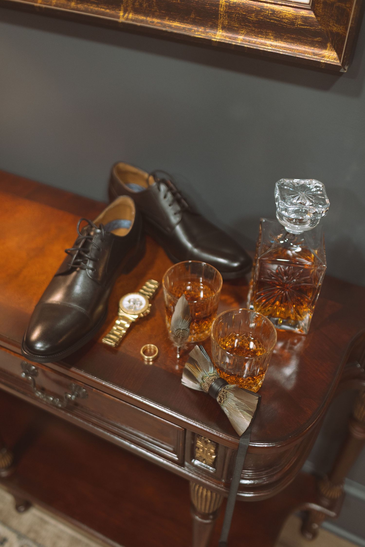 Groom accessories including black shoes, whiskey decanter, watch on mahogany table.