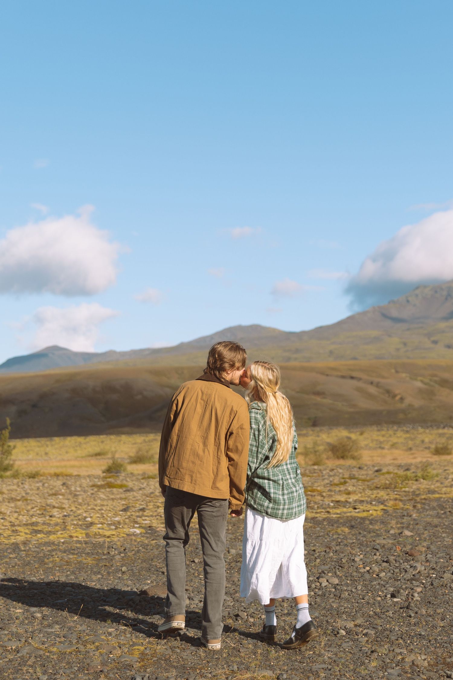 Couple walks hand in hand through a scenic volcanic landscape with mountains in background.
