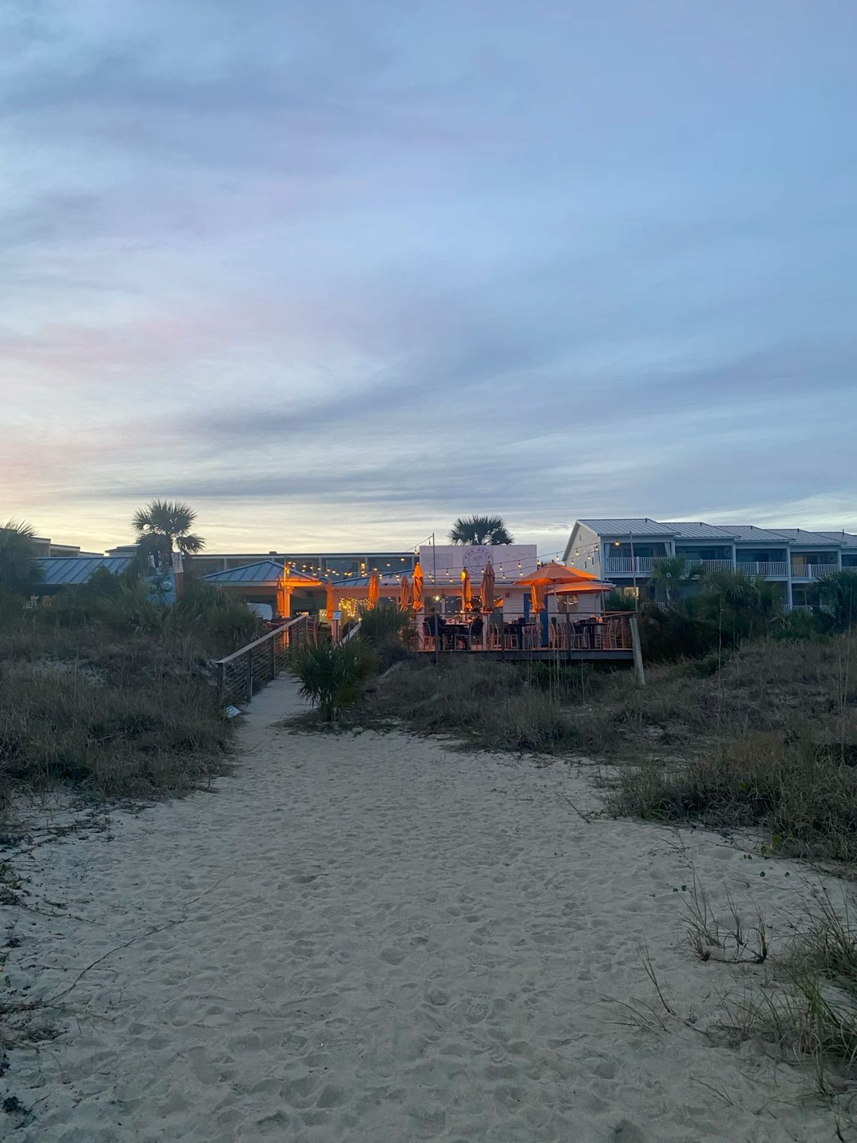 Sandy beach path at dusk leading to glowing beachside restaurant deck with palm trees.