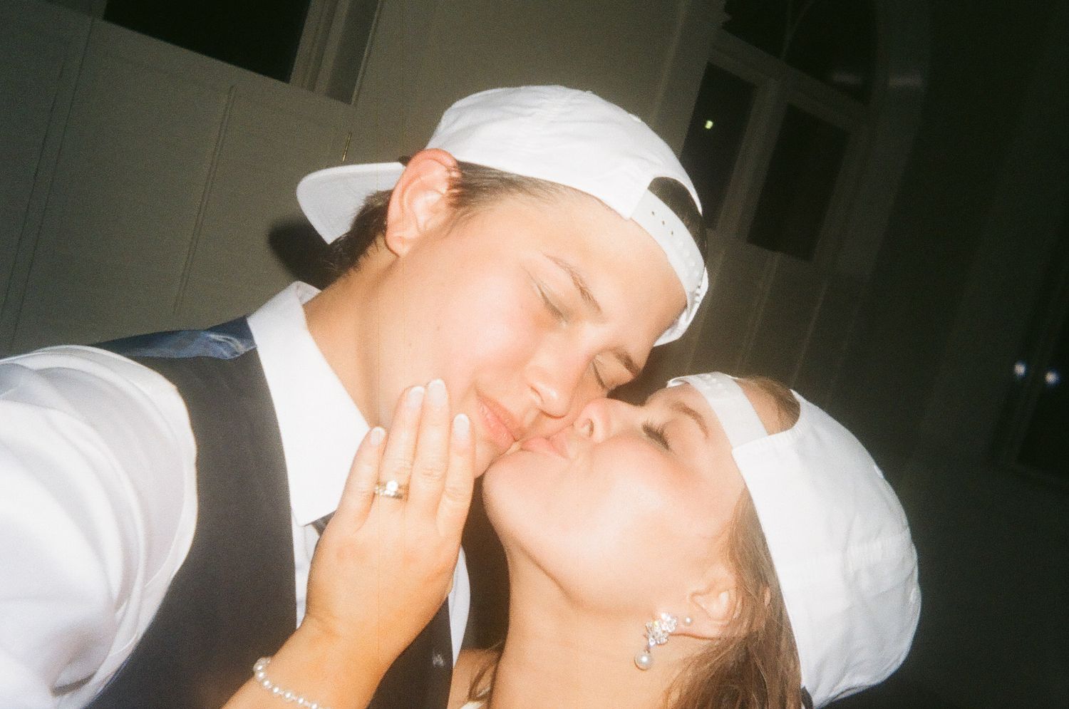 Two people wearing white caps share a romantic kiss, dressed in formal attire with jewelry visible.