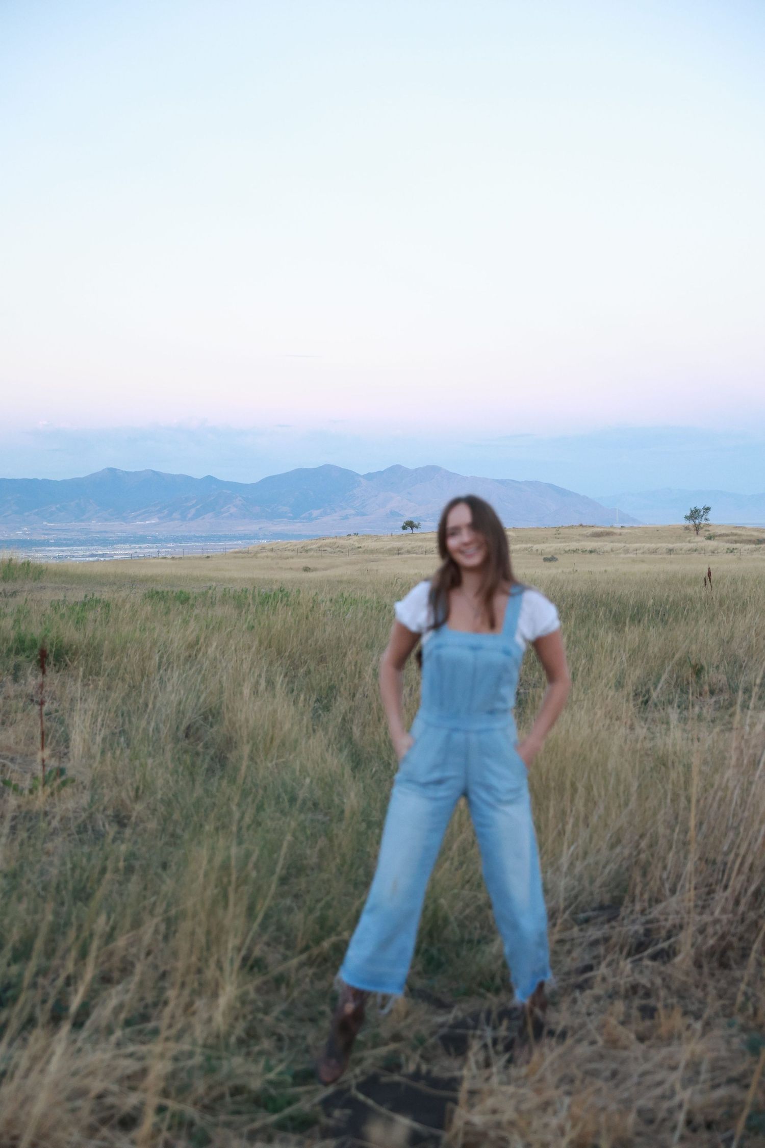 Woman in denim overalls and white tee stands in golden field with mountain backdrop at dusk.