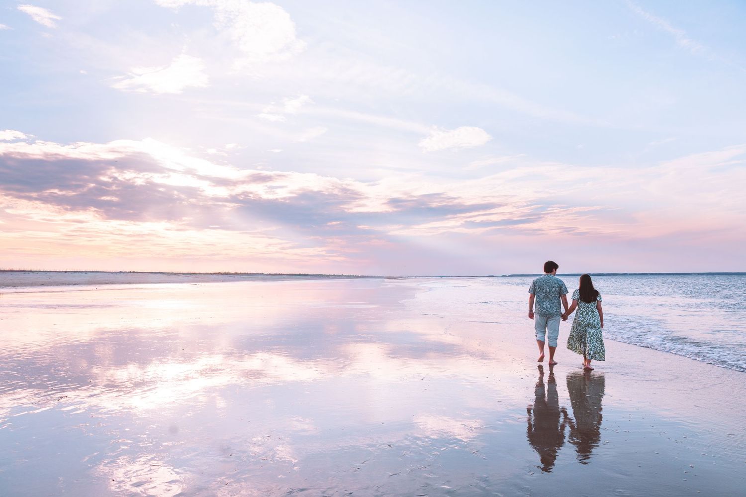 Couple holding hands walking on a reflective beach at sunset with pastel pink and blue skies.