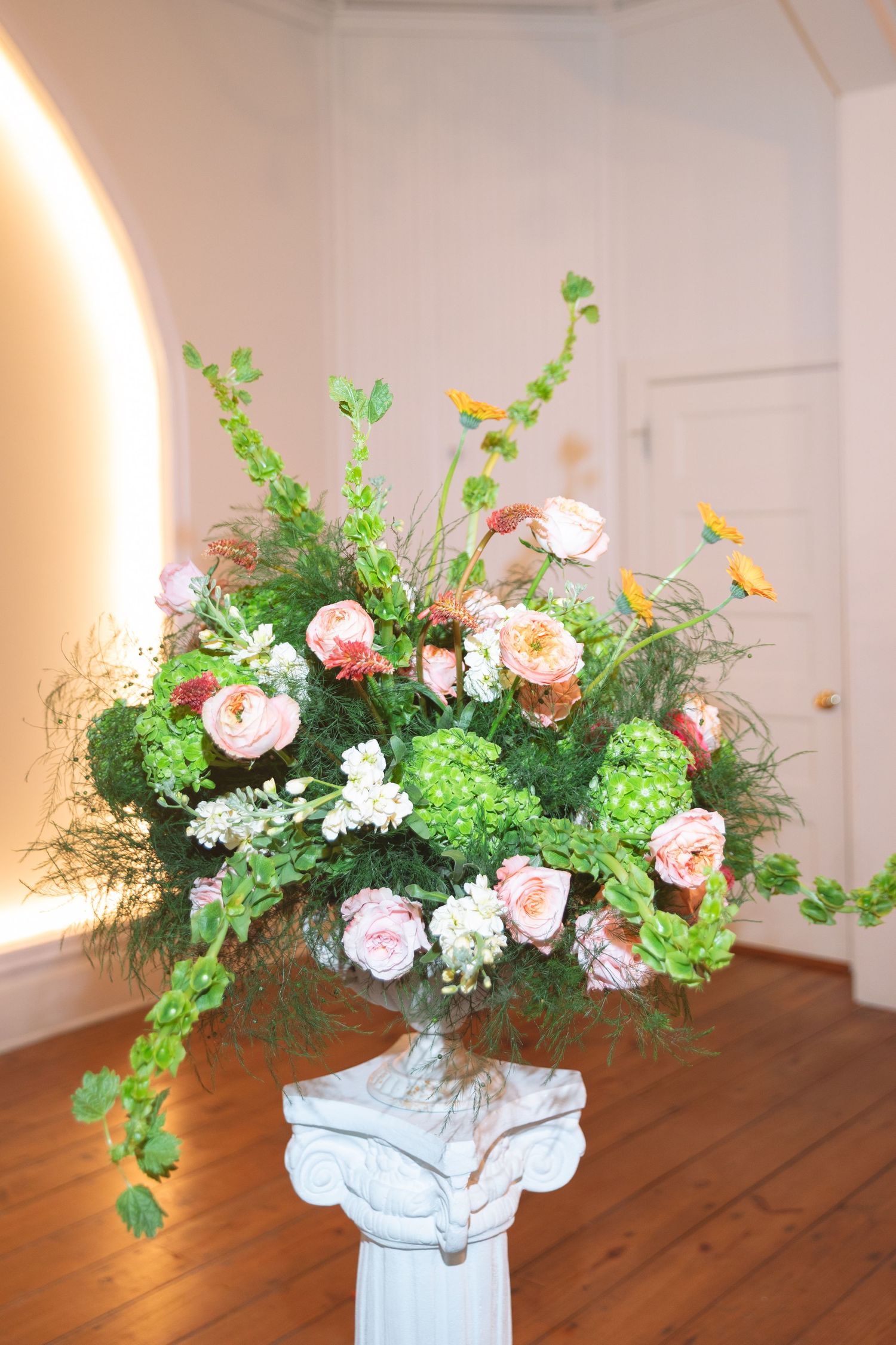 Lush floral arrangement with pink roses and greenery on a white pedestal in an elegant indoor setting.