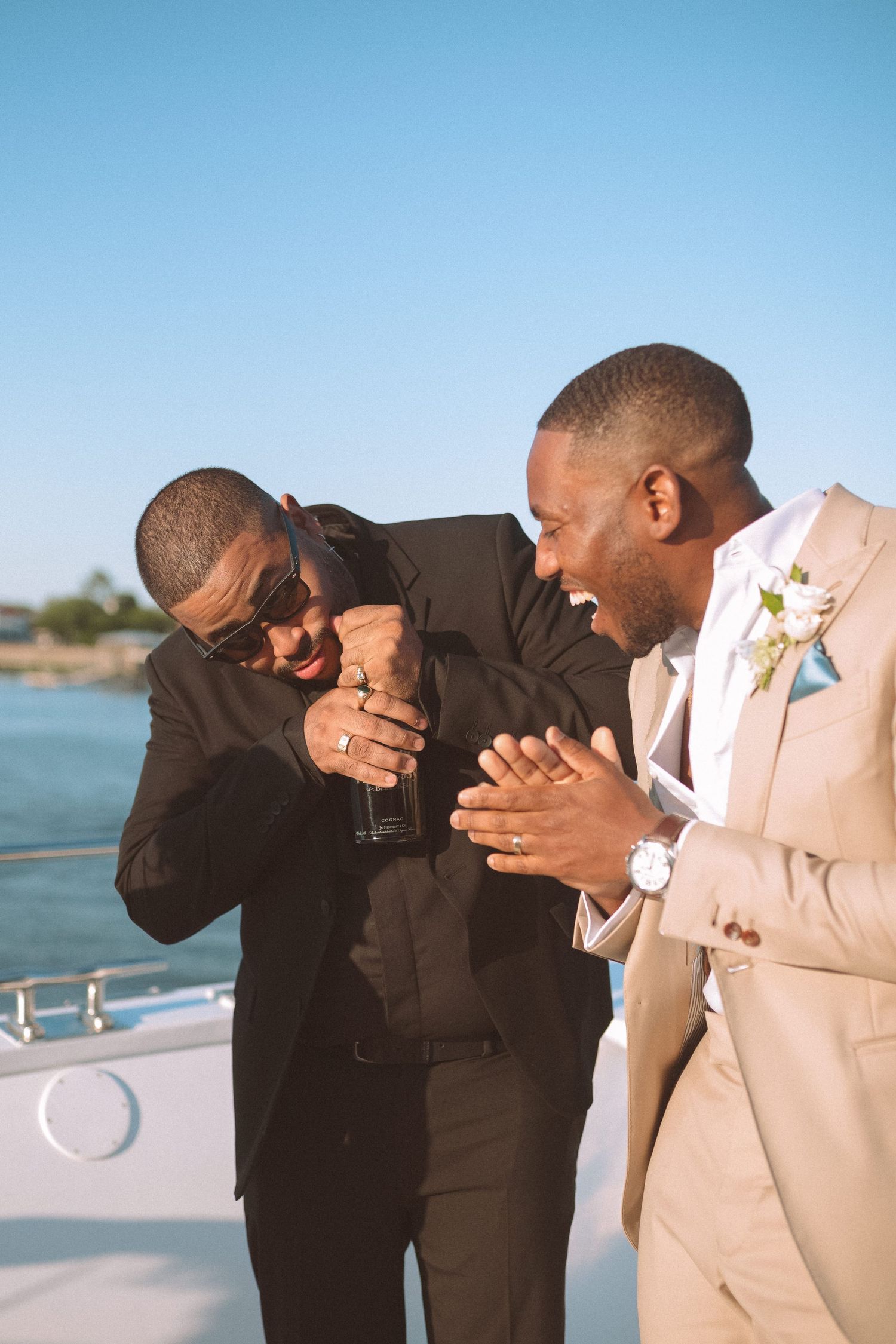 Two men in formal attire laughing together while opening champagne bottle on yacht deck.