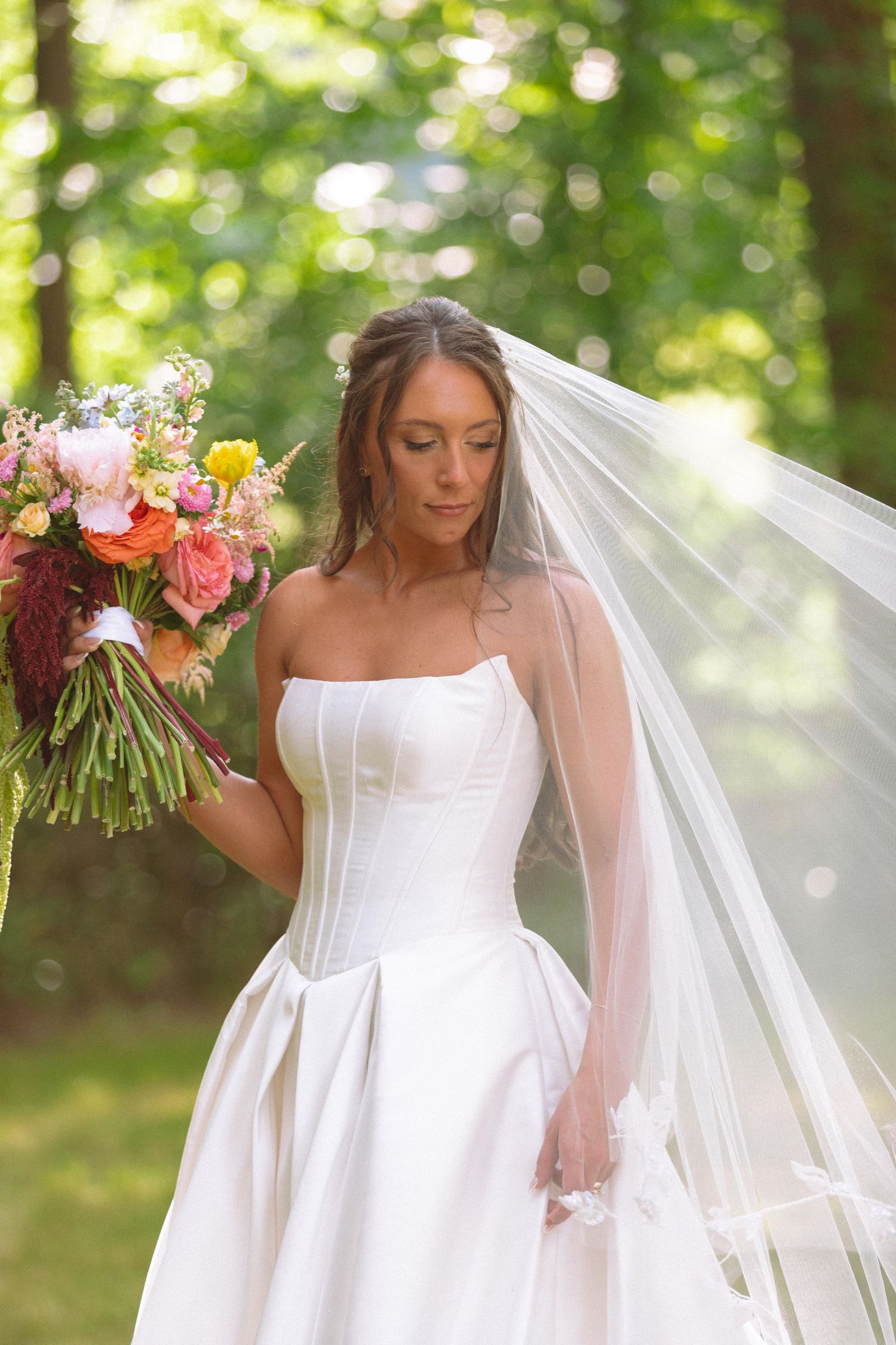 Beautiful bride in strapless corset gown holds vibrant bouquet with flowing veil in garden.