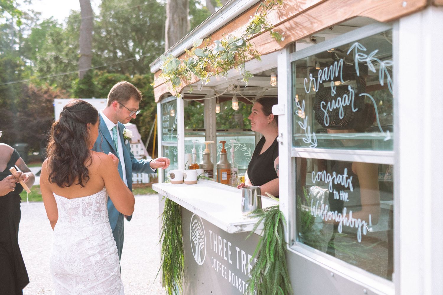 Bride and groom visit rustic Three Trees bar truck at outdoor wedding reception with greenery decor.