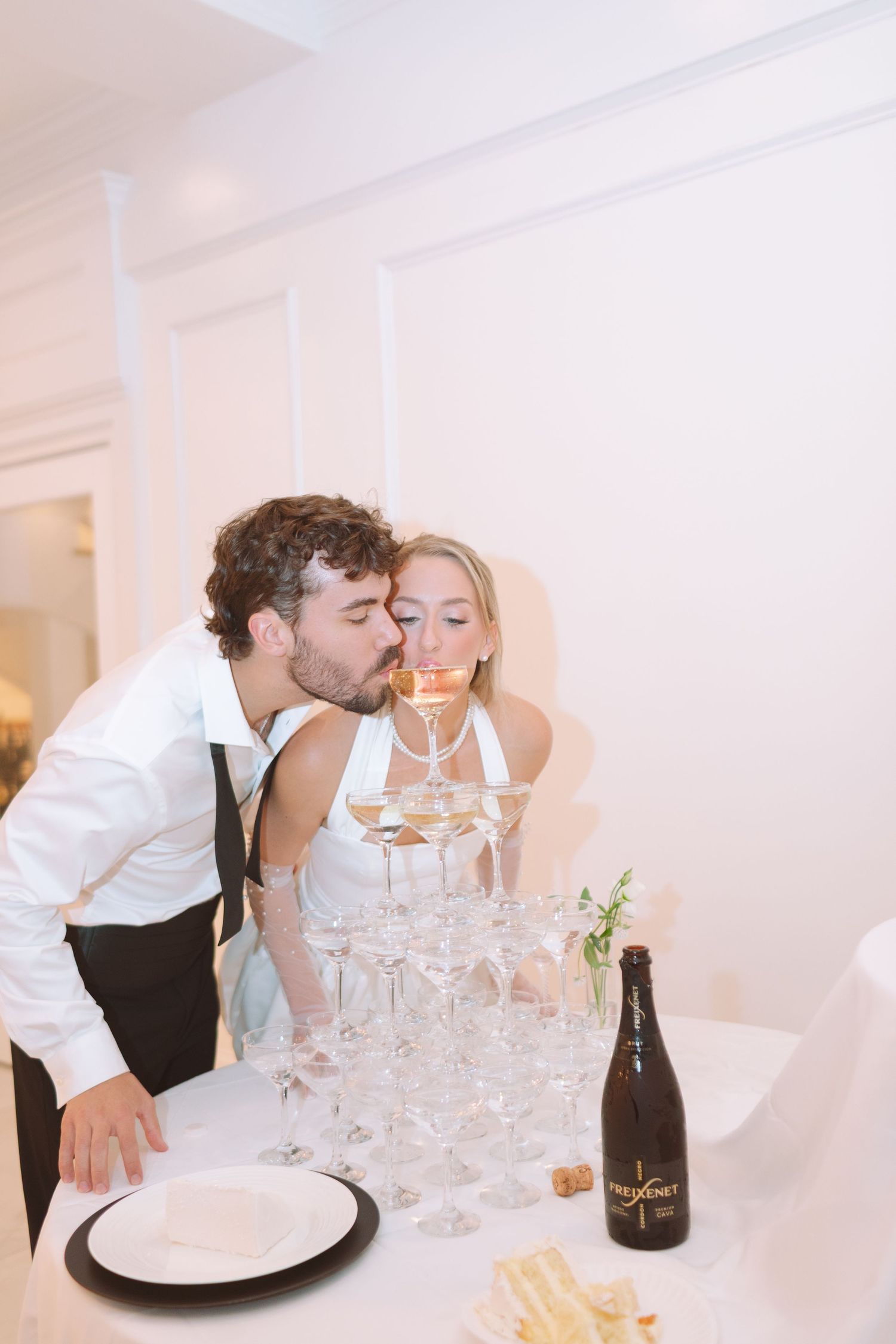 Groom kissing bride on cheek beside champagne tower at wedding reception.