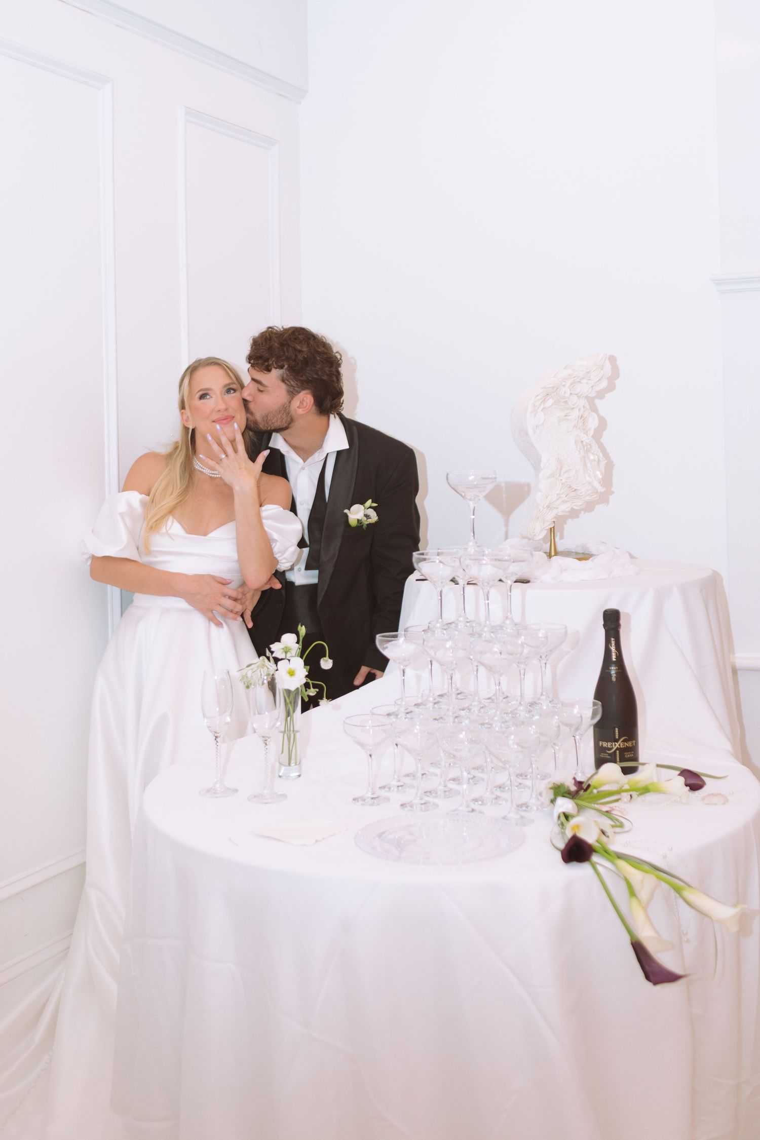 Bride and groom posing beside a champagne tower with a bottle and floral bouquet at their wedding reception.
