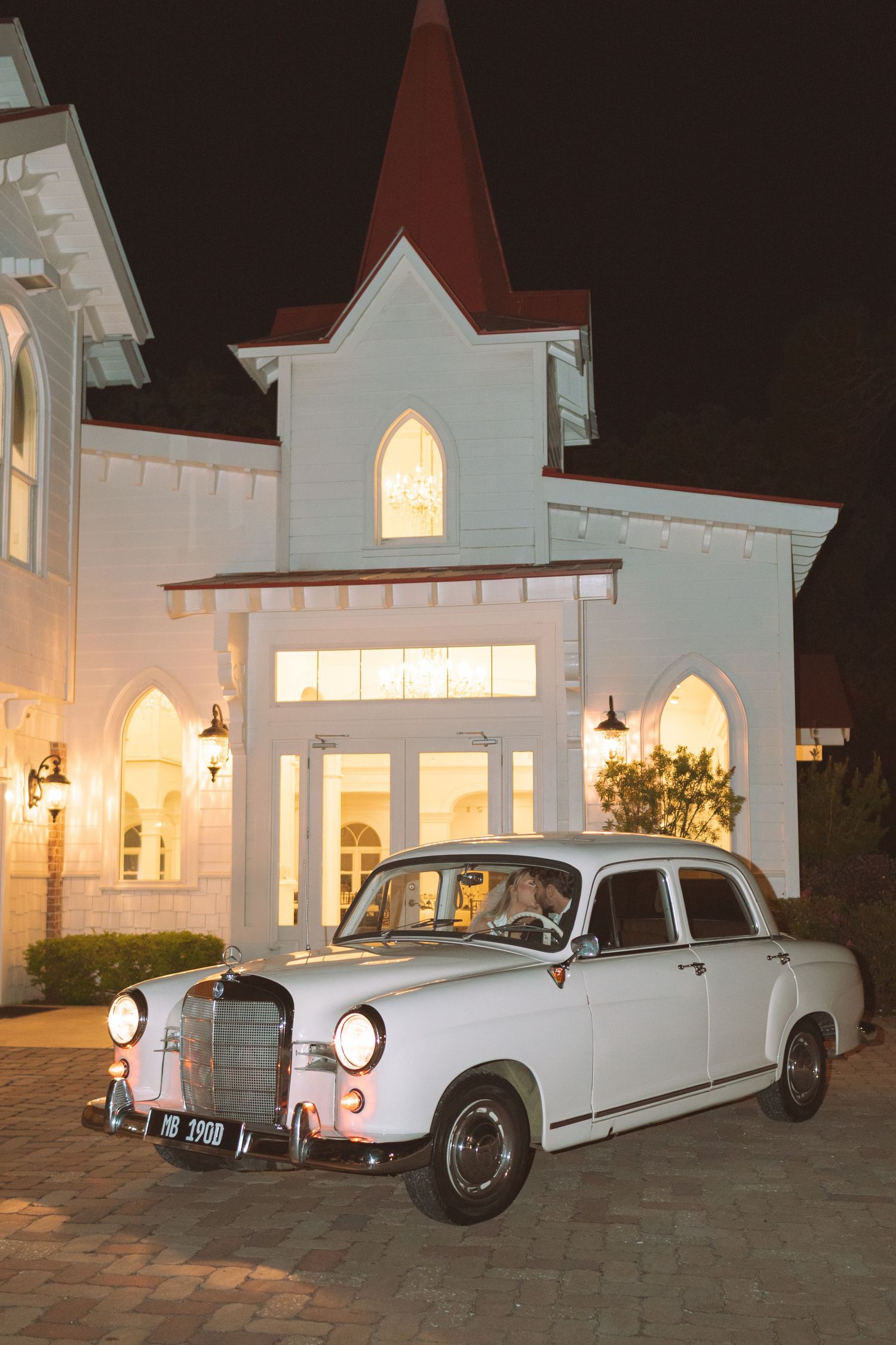 Classic vintage white Mercedes parked outside illuminated white chapel wedding venue at night.