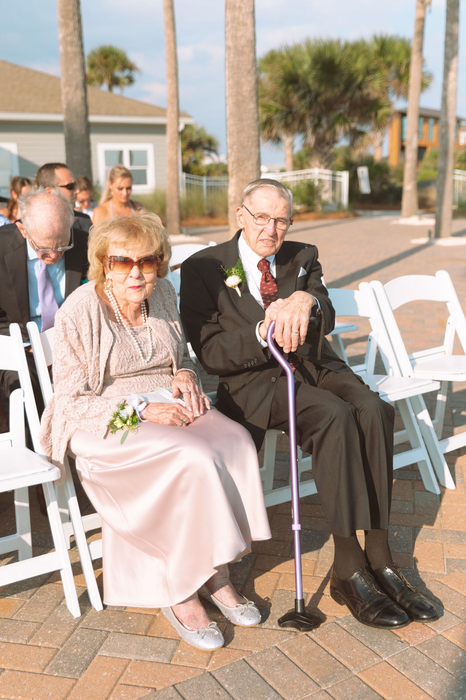 Elderly couple in formal attire sitting together at an outdoor wedding ceremony with palm trees behind.