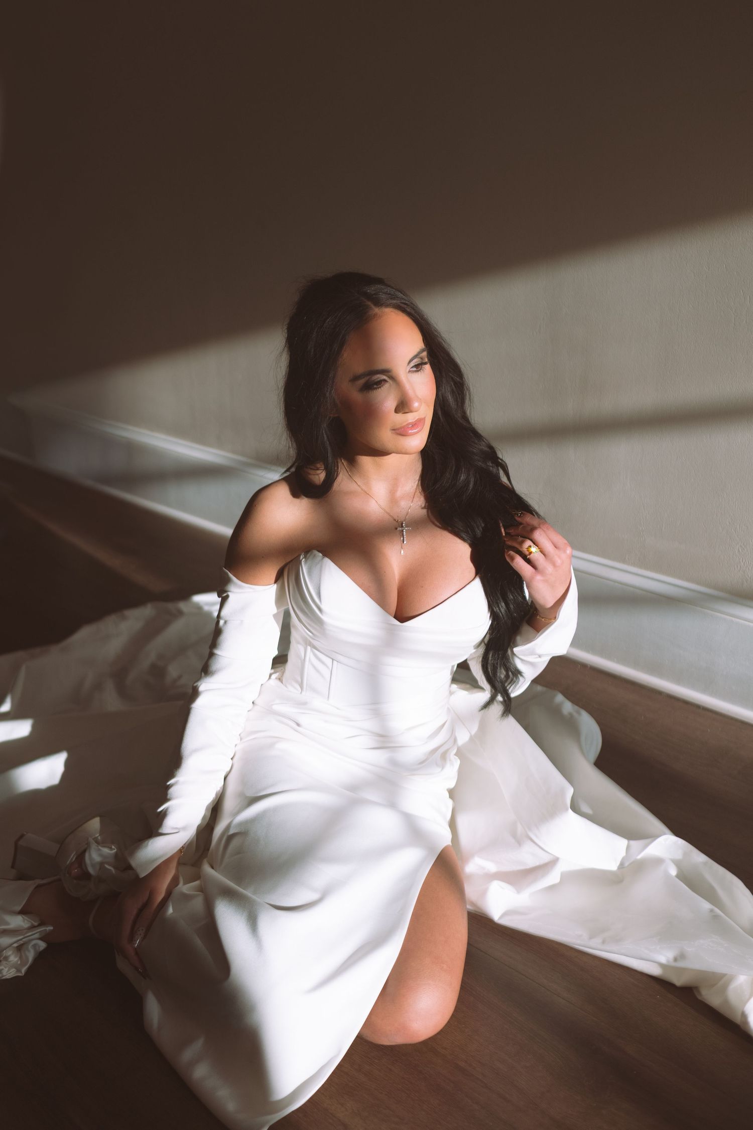 Elegant bride in a white off-shoulder satin gown sits on stairs in soft natural light.