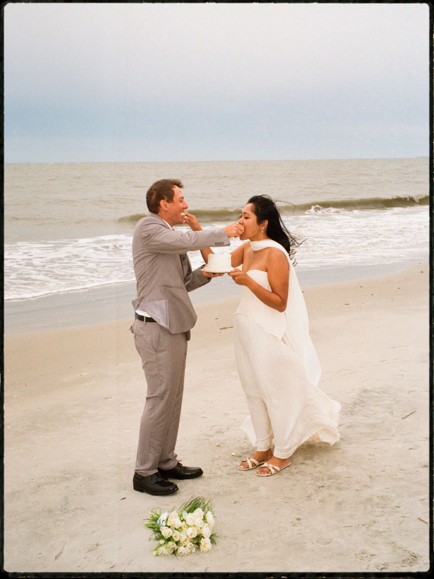 Bride and groom sharing wedding cake on a sandy beach with ocean waves in the background.