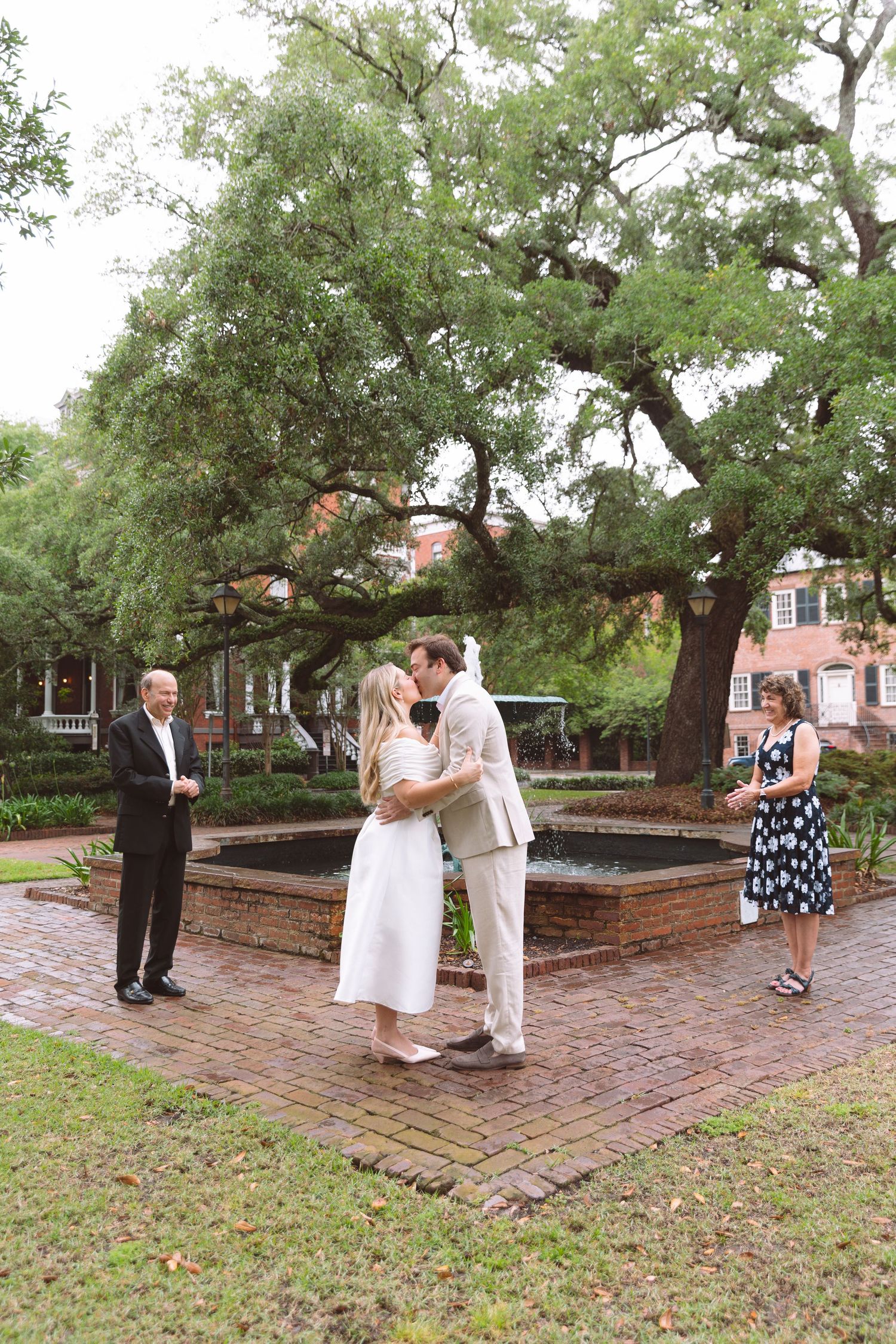 Couple shares first kiss at outdoor elopement ceremony by brick fountain with officiant and witness present.