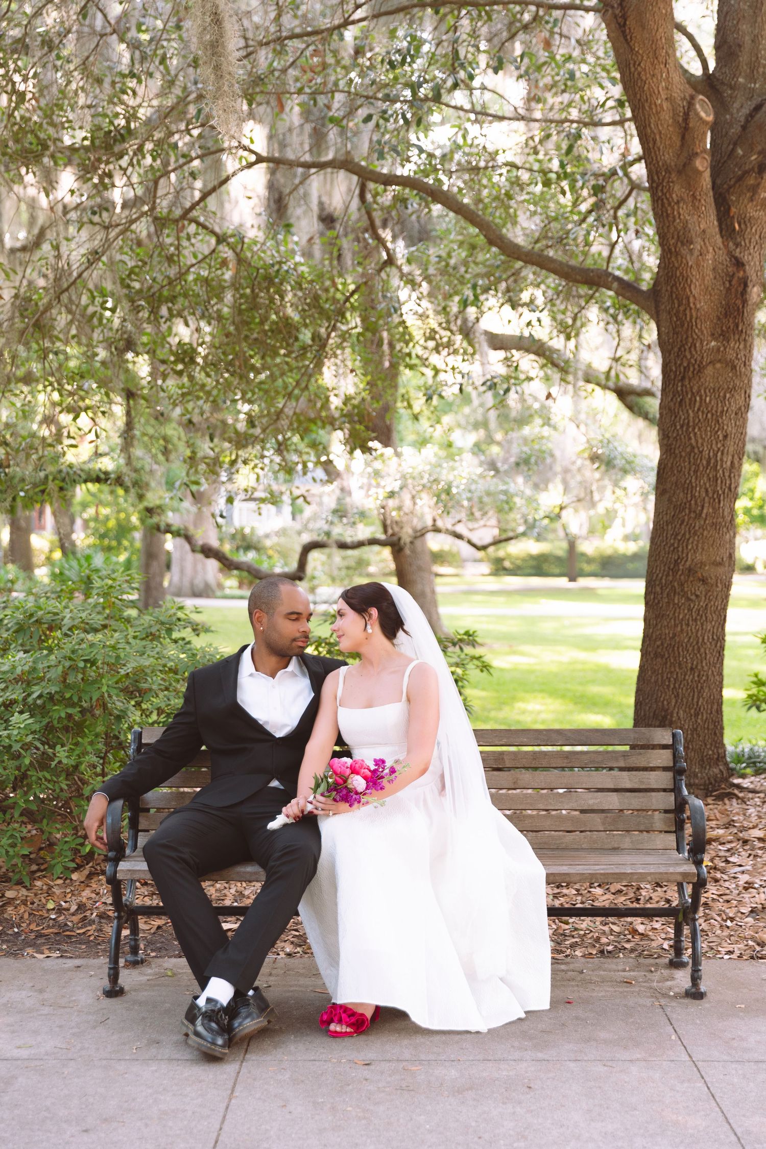 Interracial couple in wedding attire sitting on a park bench, sharing a romantic moment surrounded by trees.