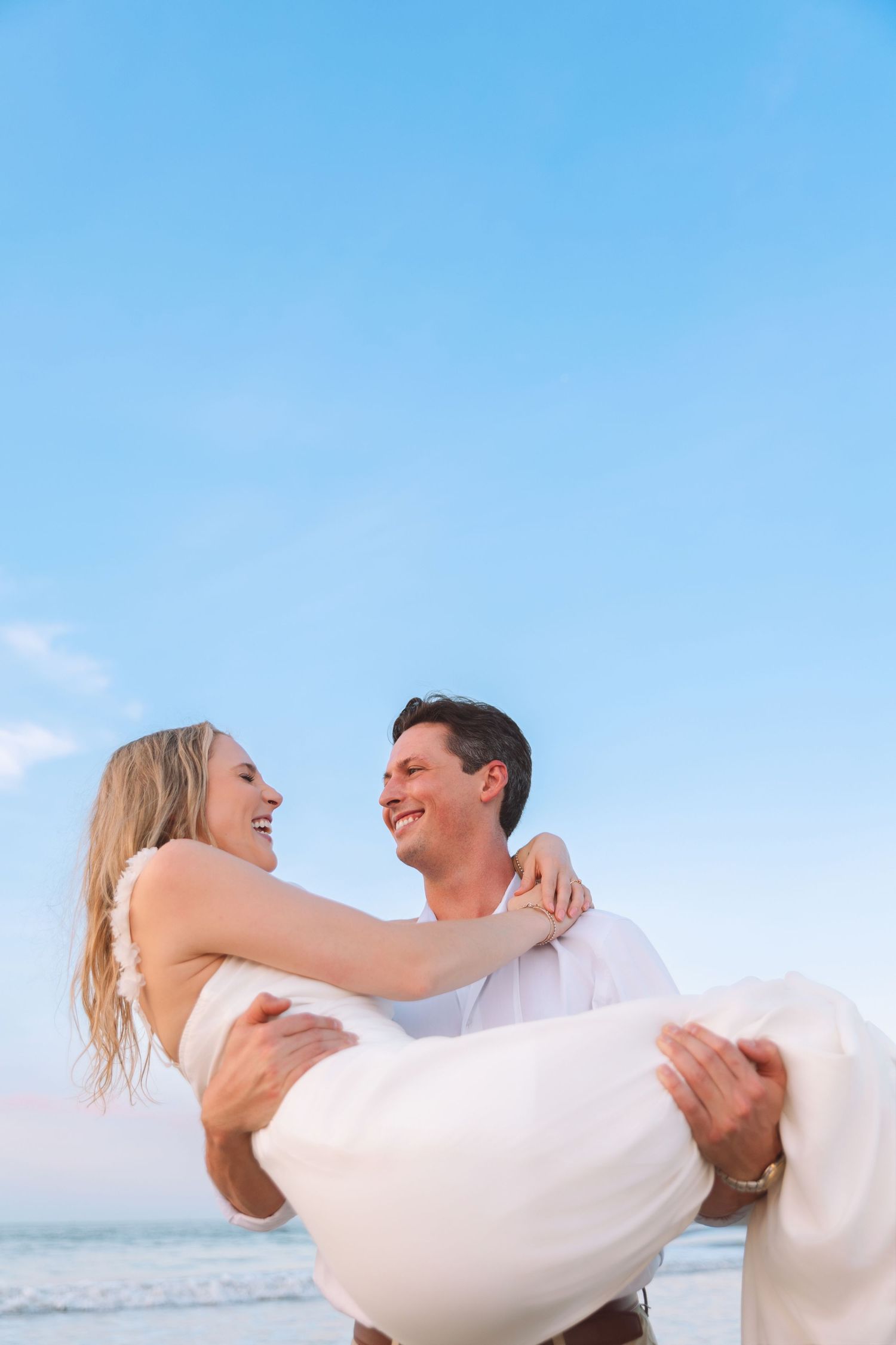 Groom carrying bride in white dress on sunny beach, both laughing joyfully together.