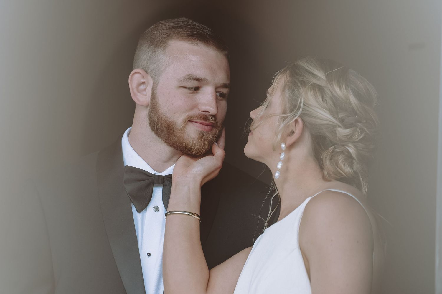 Bride tenderly touches bearded grooms face during intimate wedding portrait in soft light.