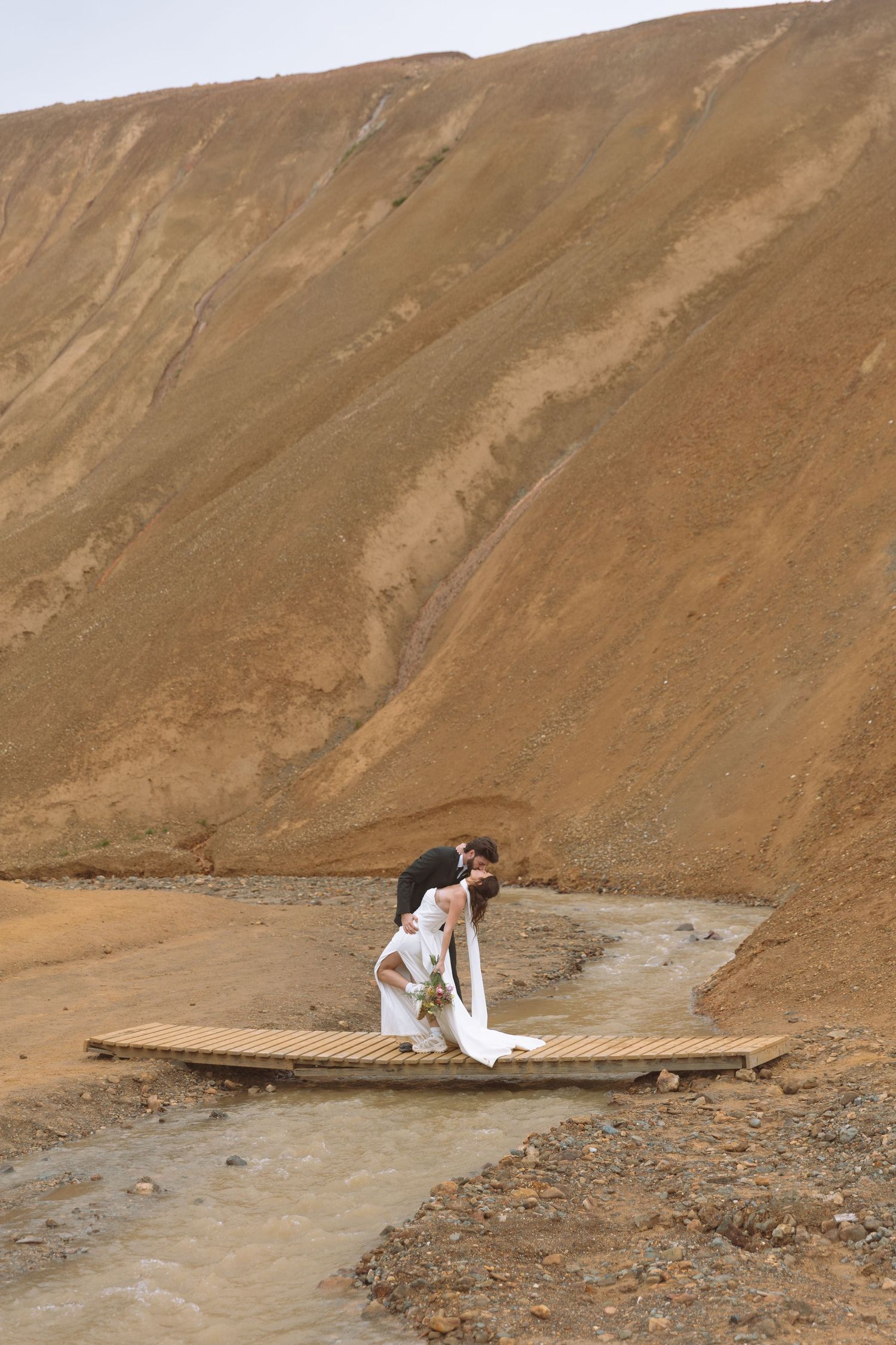Groom dips bride on a rustic wooden bridge with Iceland's rugged rhyolite mountains in the background.