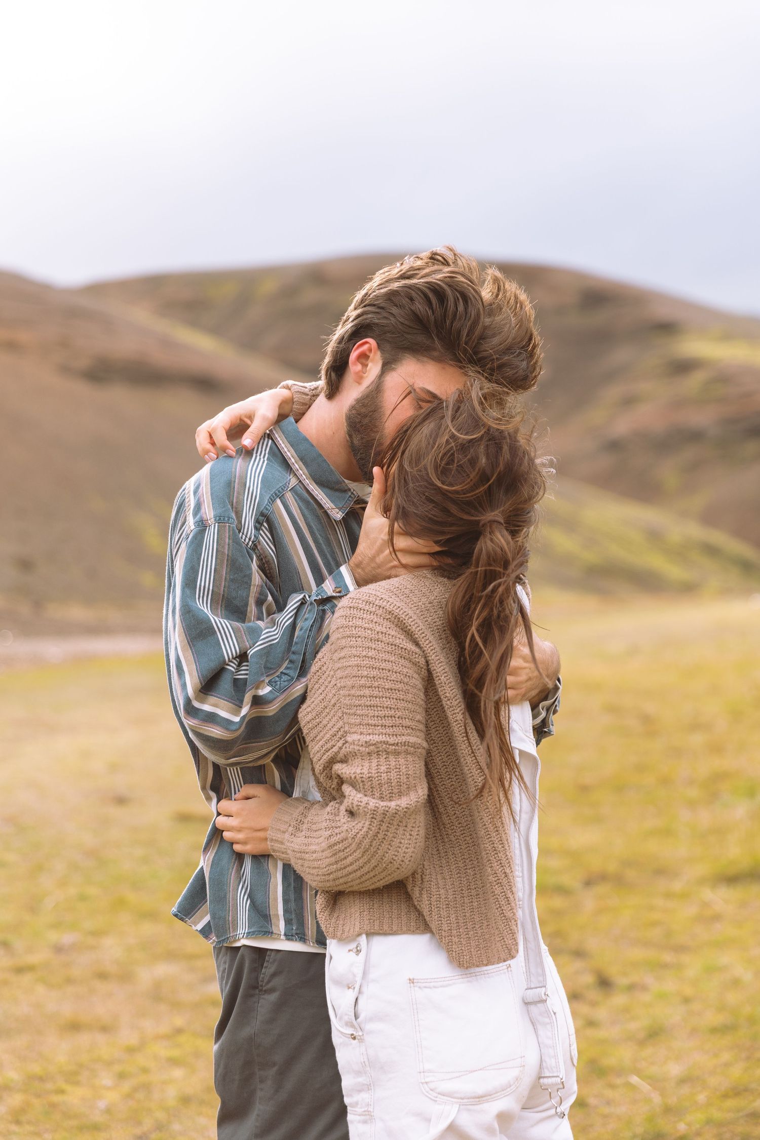 Couple hugging closely in a windy green mountain meadow under cloudy skies.