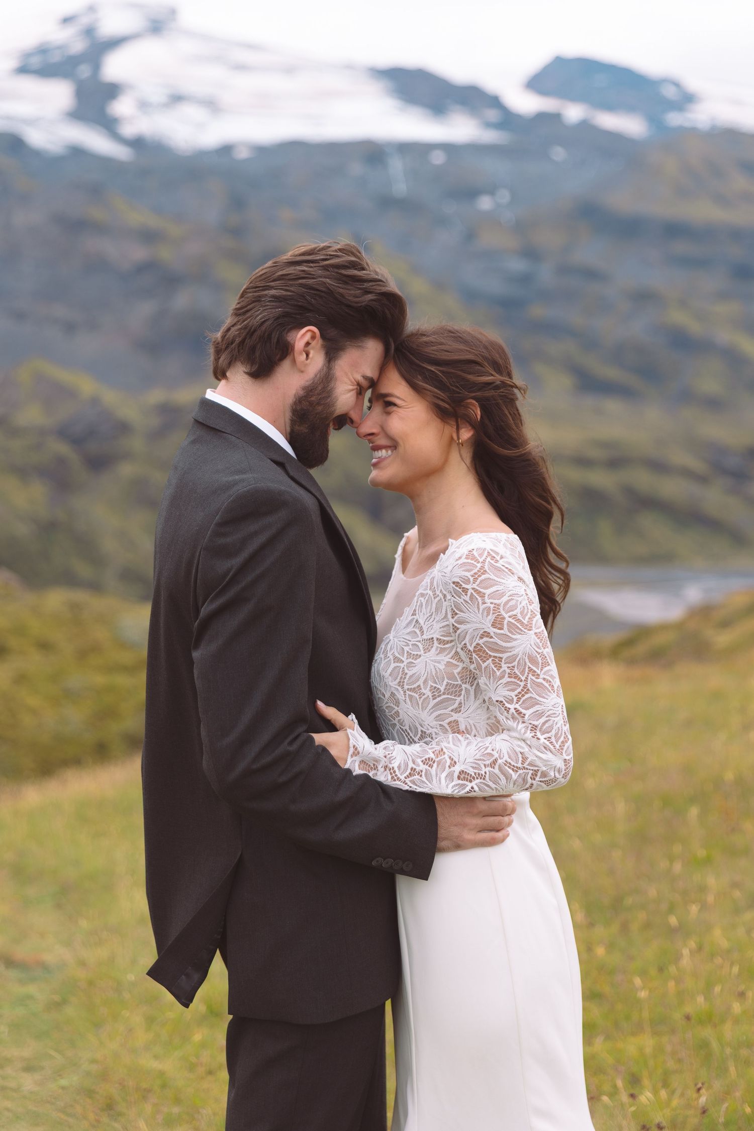 Bride and groom embrace forehead to forehead in a romantic outdoor wedding portrait with snow-capped mountains behind.