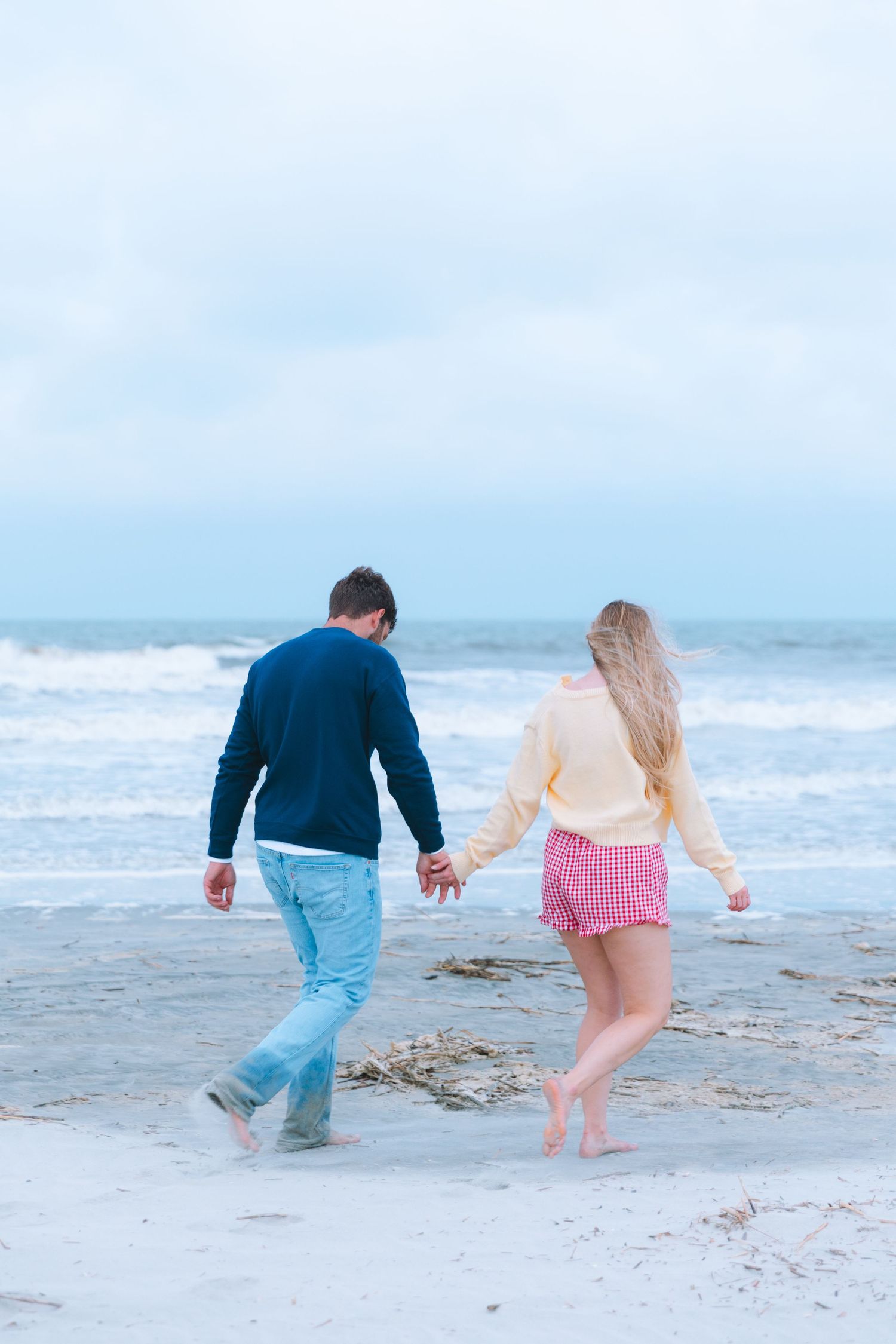 Couple holding hands walking barefoot on a sandy beach toward ocean waves on a cloudy day.