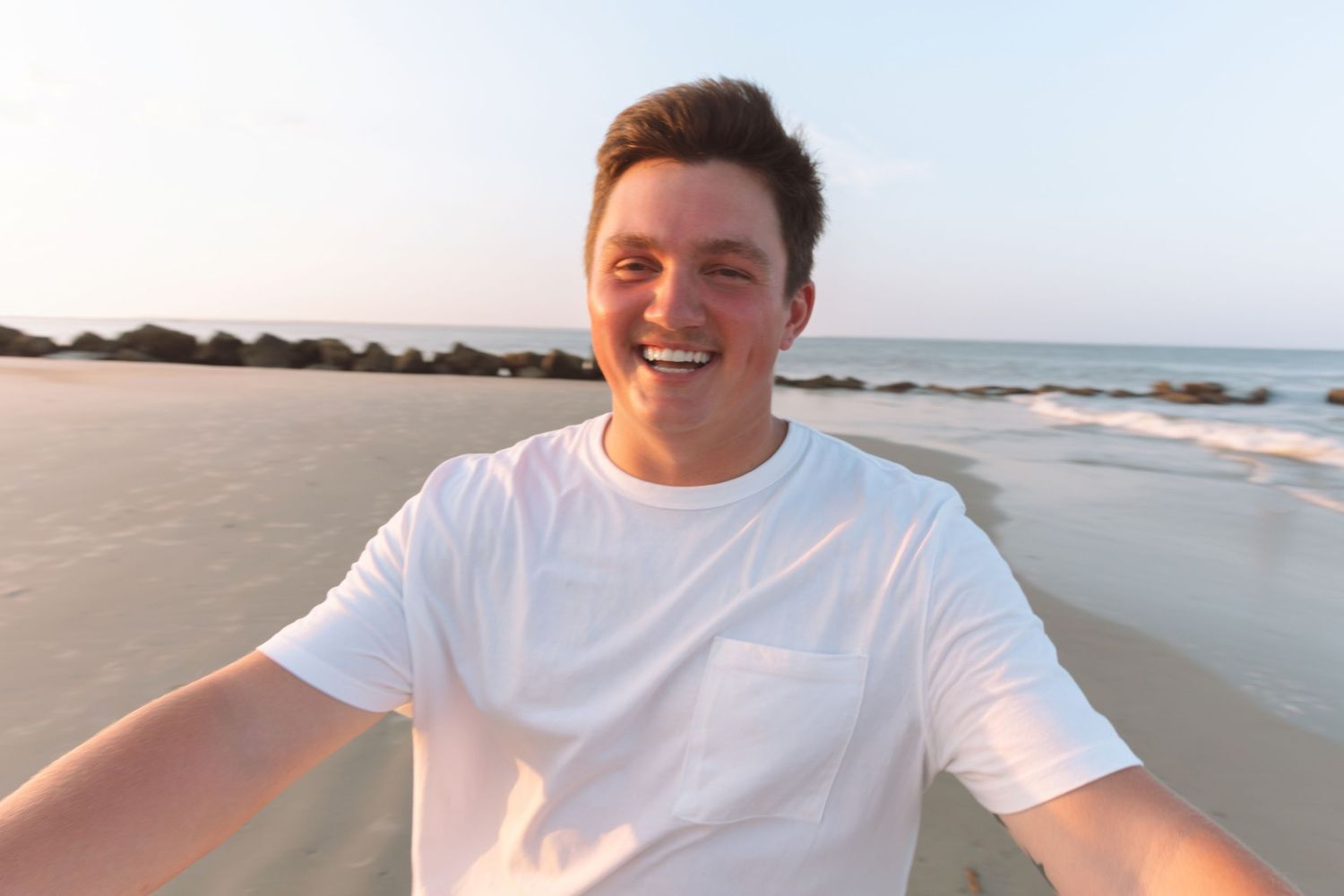Smiling young man in white t-shirt taking a selfie on a sunny beach with ocean in the background.