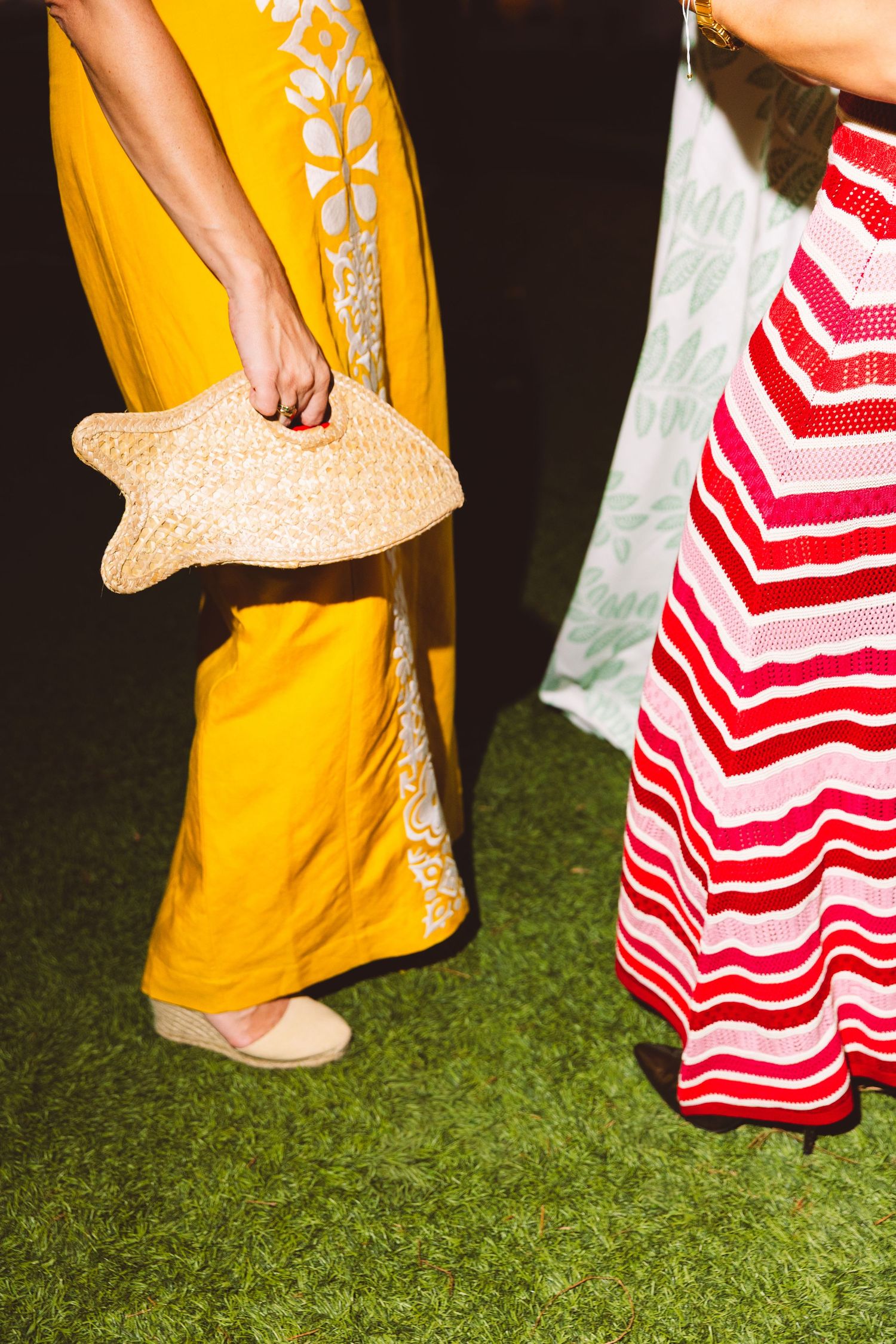 Two women in colorful maxi dresses, one yellow with floral print holding a straw hat, on green grass.