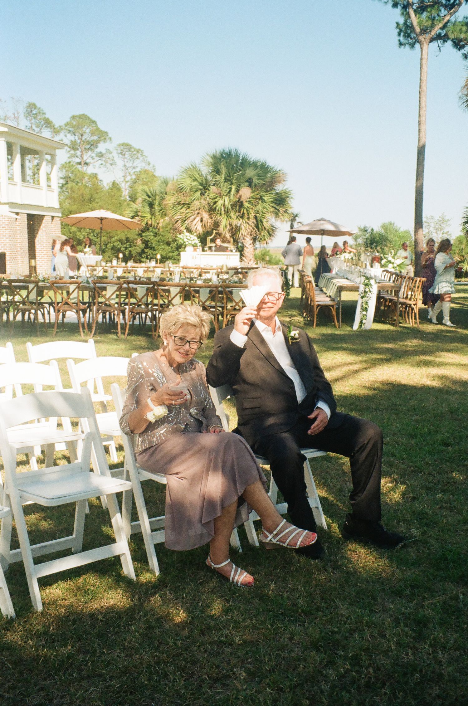 Elderly couple fans themselves with programs while seated in white chairs at an outdoor garden wedding ceremony.