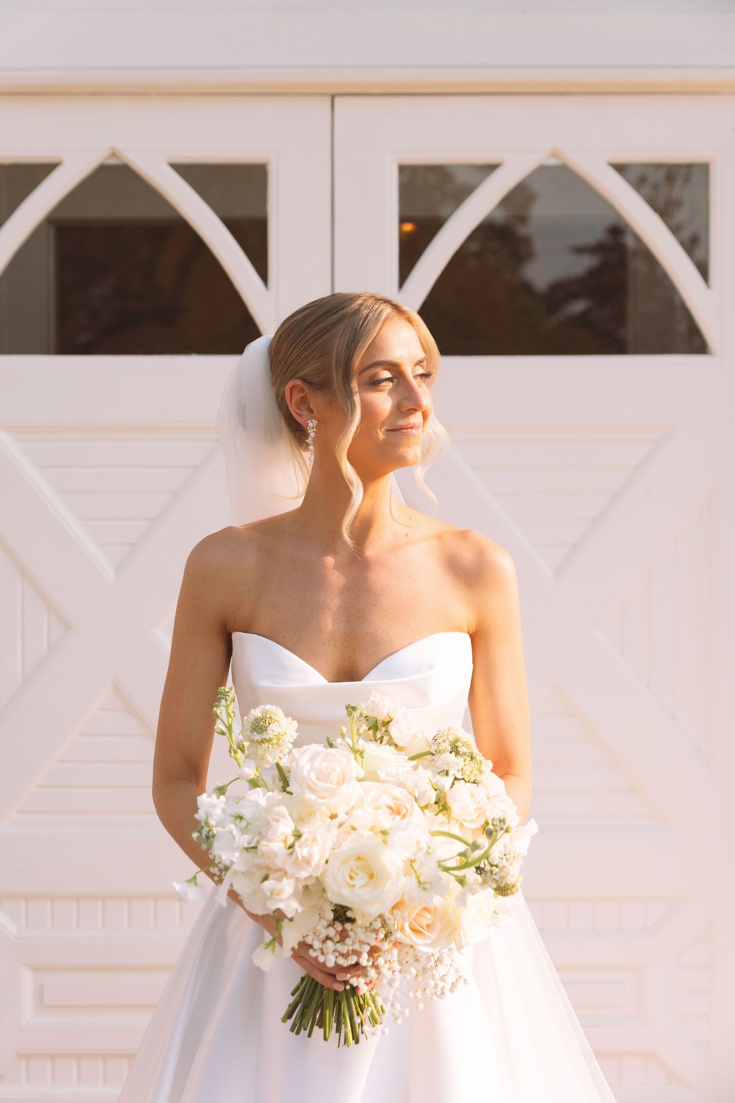 Smiling bride in strapless white gown holding lush white rose bouquet against decorative white doors.