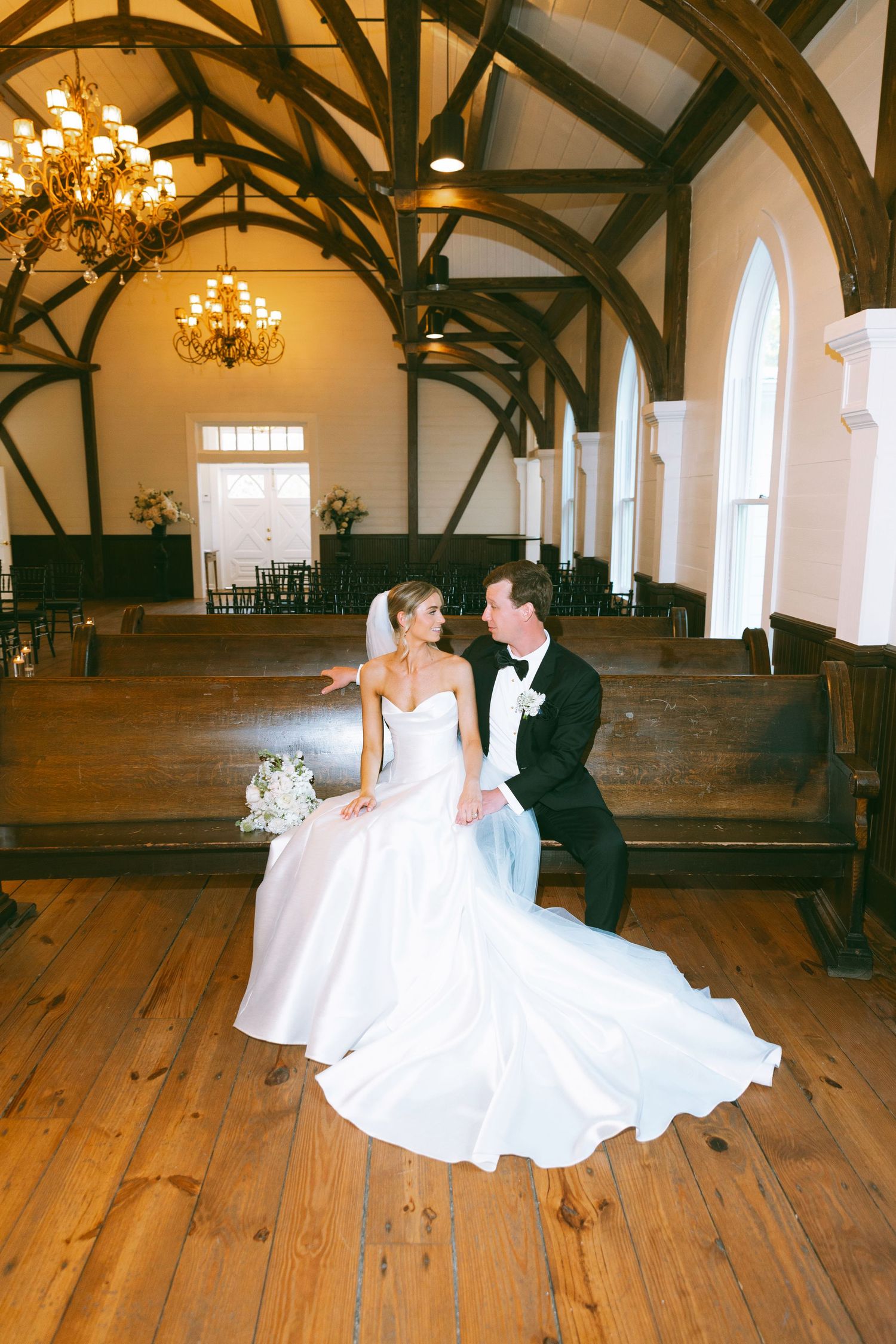 Bride and groom sit intimately on church pew inside rustic white chapel with wooden beams and chandeliers.