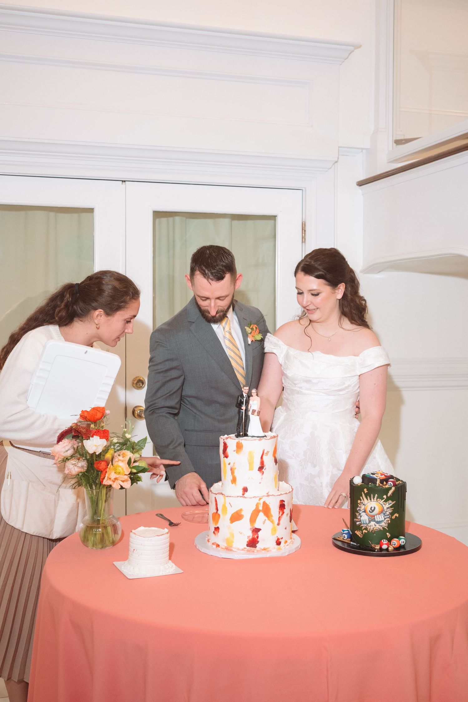Bride and groom cutting a two-tier wedding cake with autumn leaf design, assisted by a wedding planner at a reception.