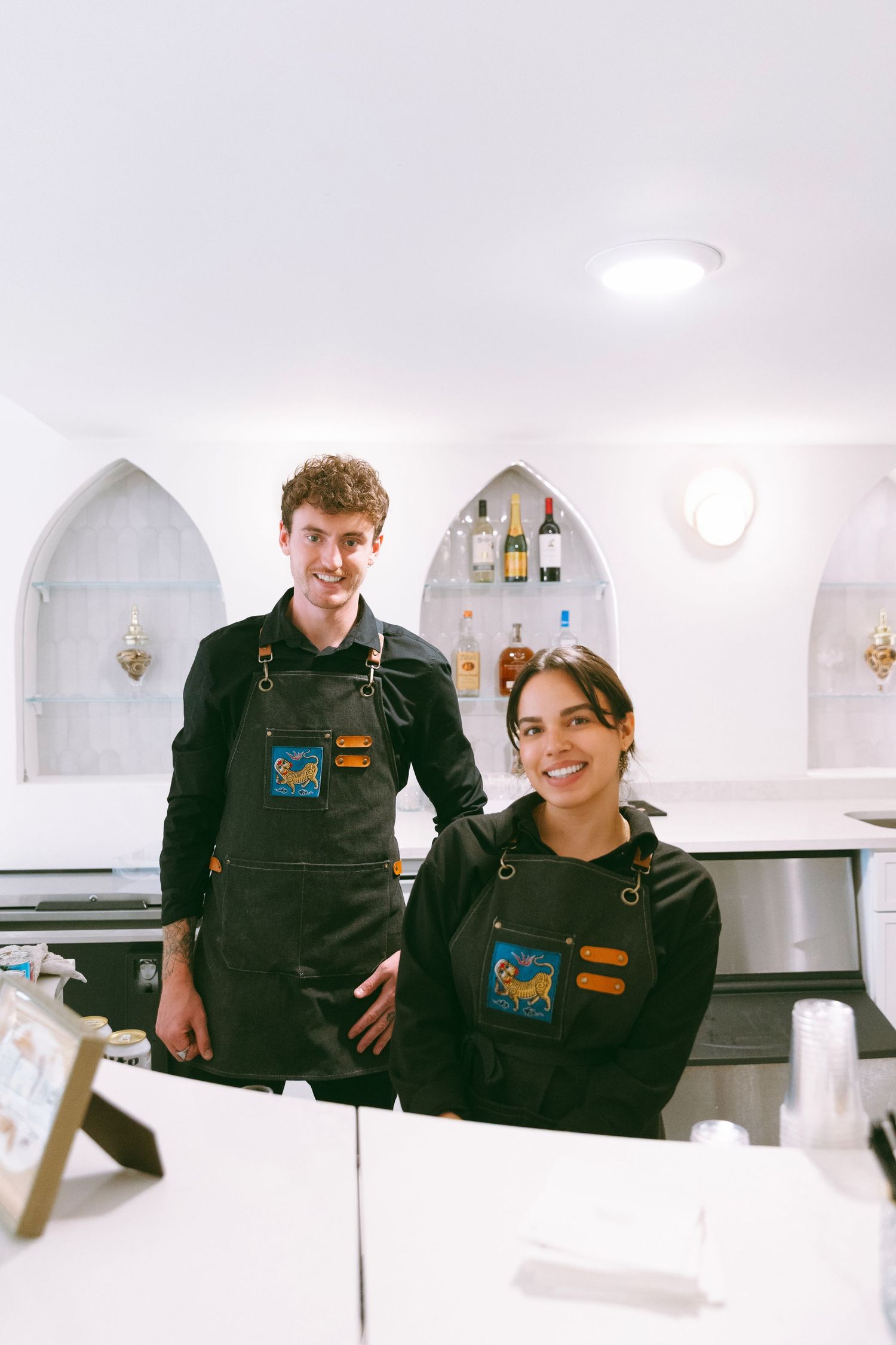 Two smiling bartenders in matching dark aprons posing behind a white wedding bar.