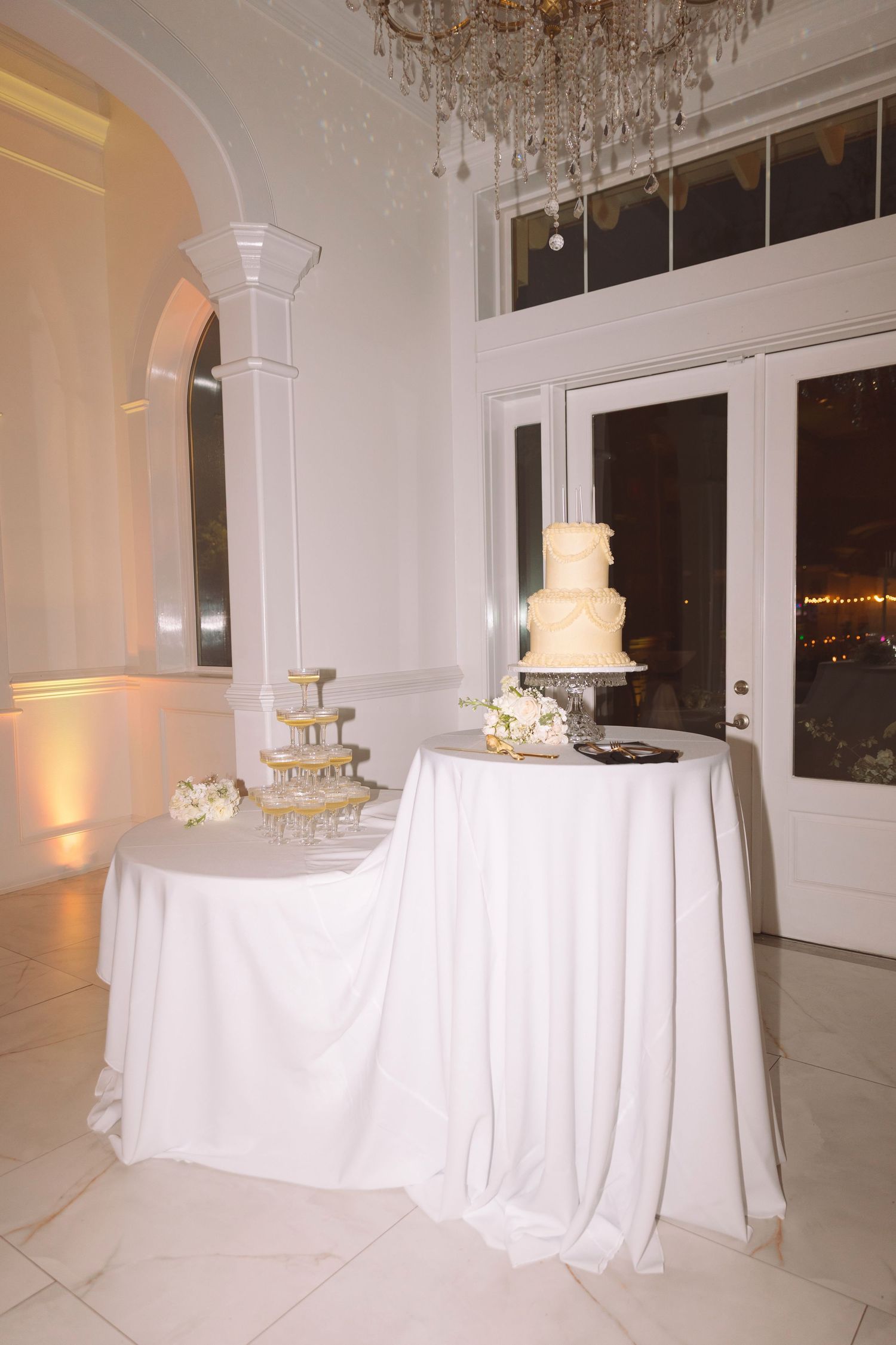 Elegant white wedding cake and champagne tower display on draped tables in a grand ballroom.