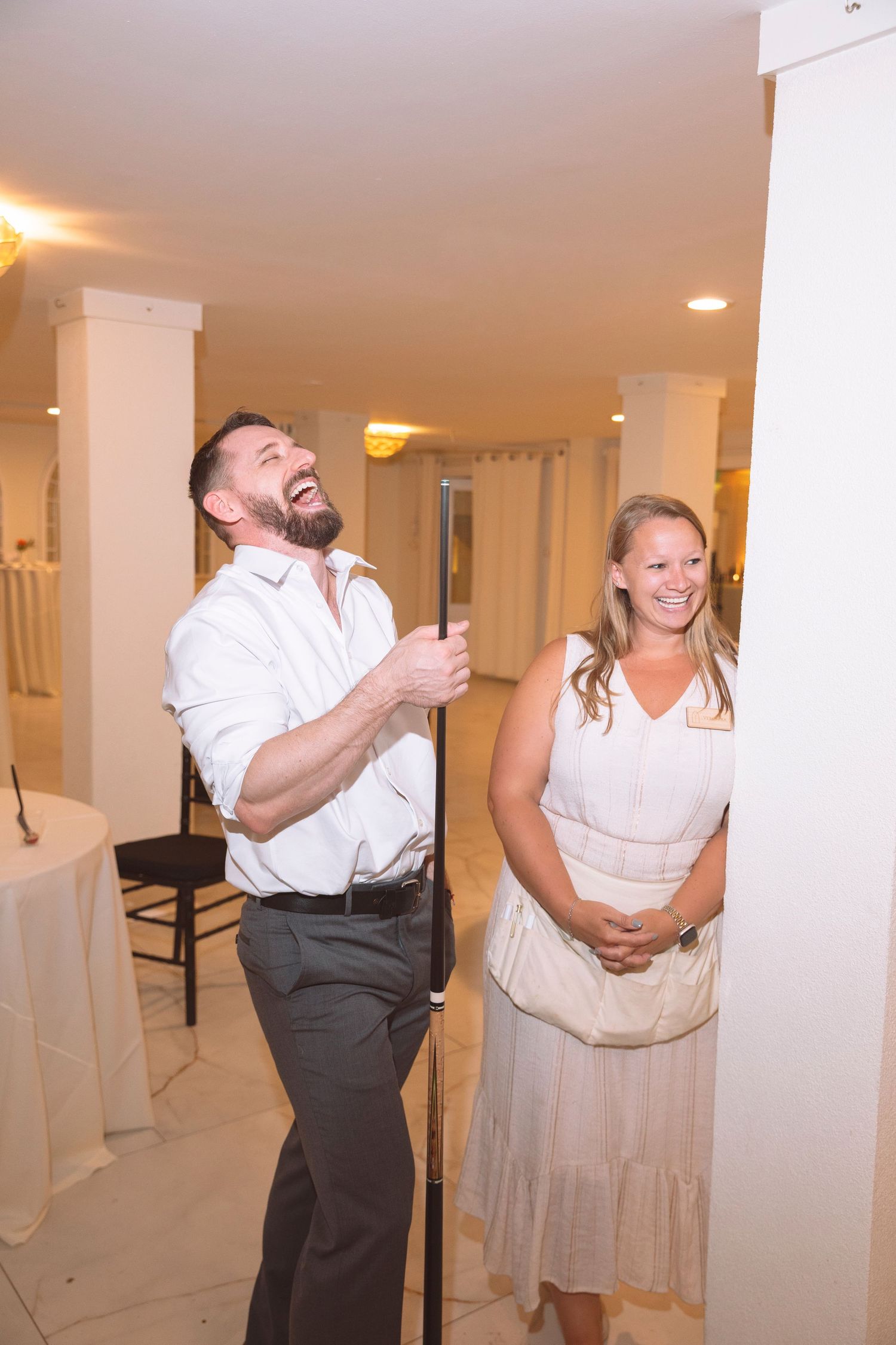 Man laughing while holding a pole next to a smiling woman in a elegant white venue.