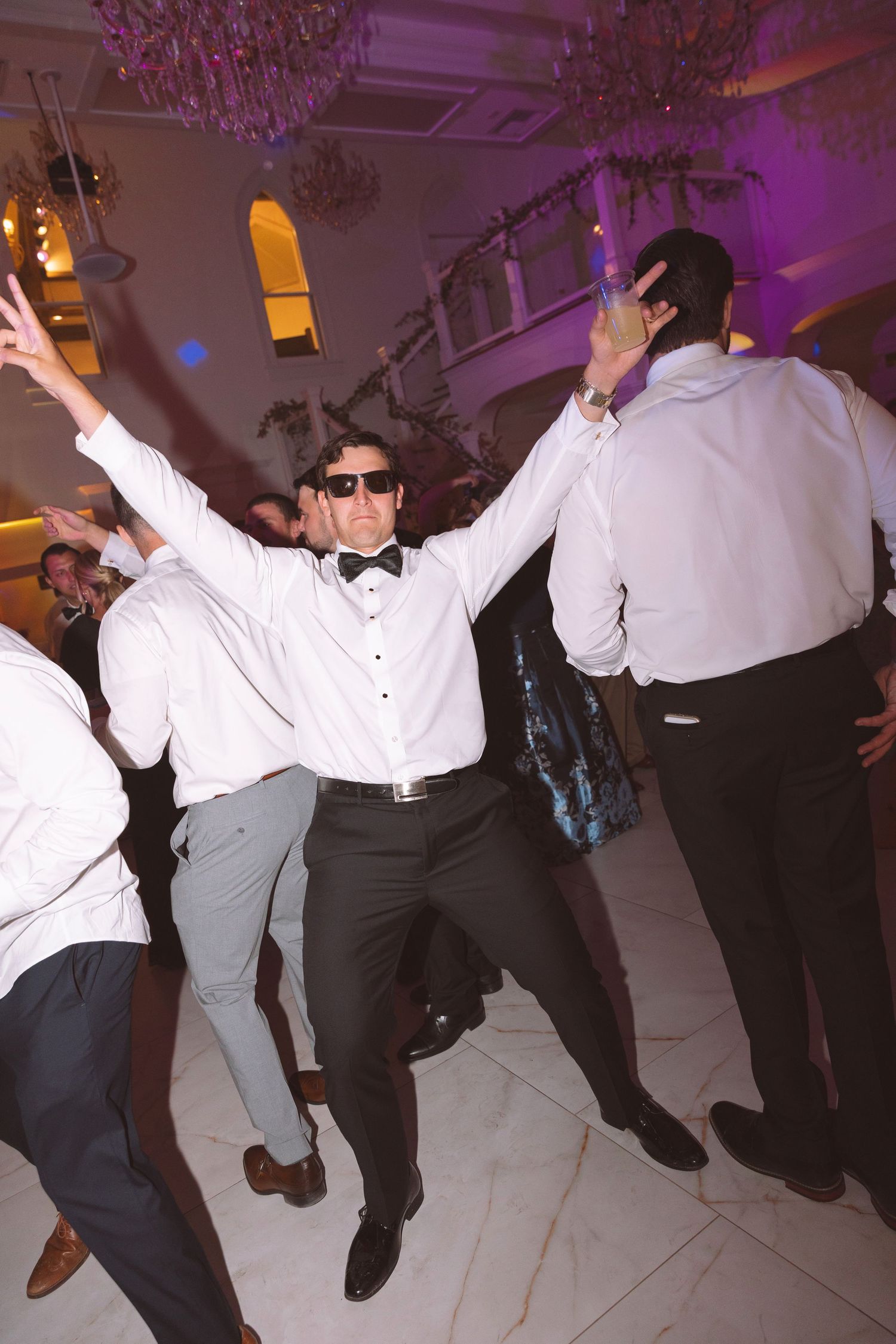 Groom in sunglasses and bow tie dancing energetically with guests at a wedding reception.