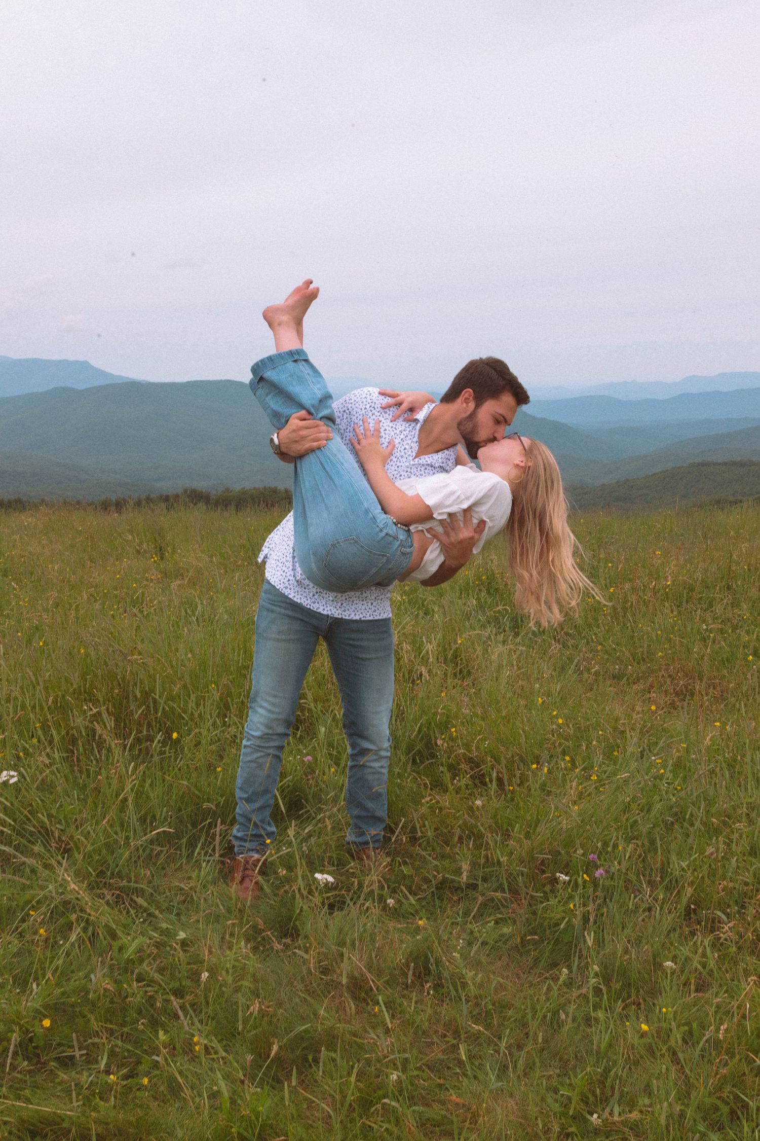 Romantic couple embracing and kissing in a green mountain meadow under cloudy skies.