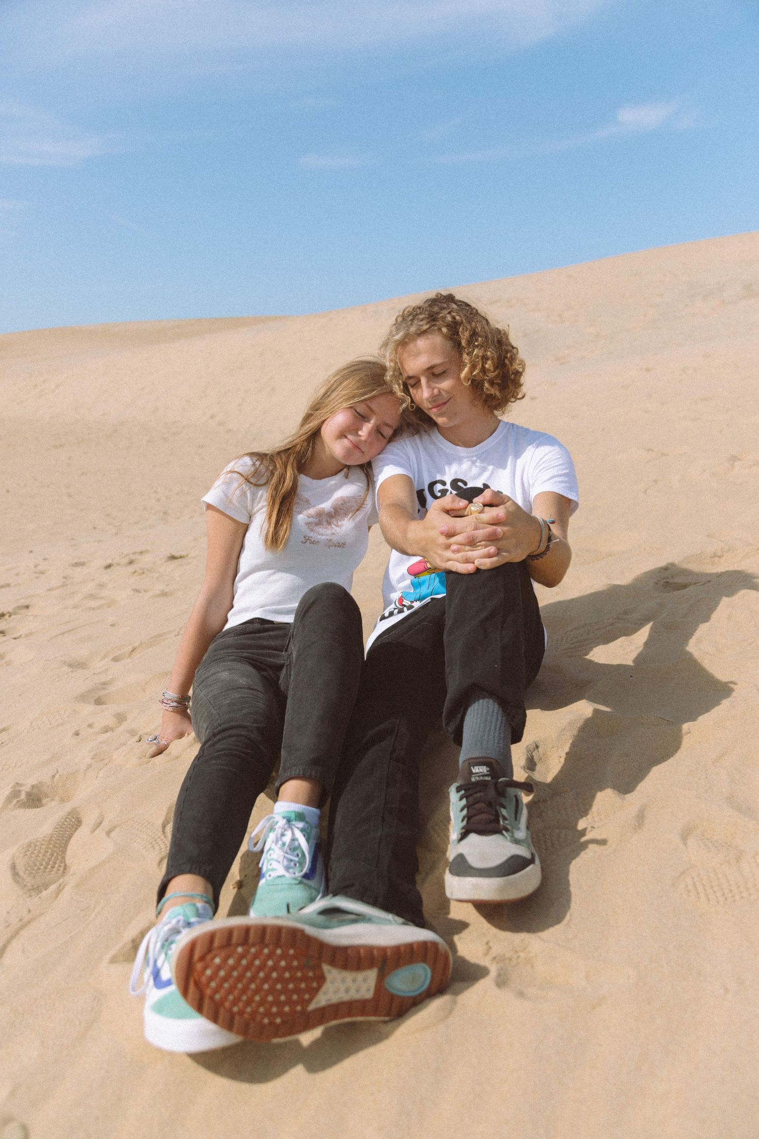 Young couple sitting together on a sandy dune, leaning on each other under blue sky.