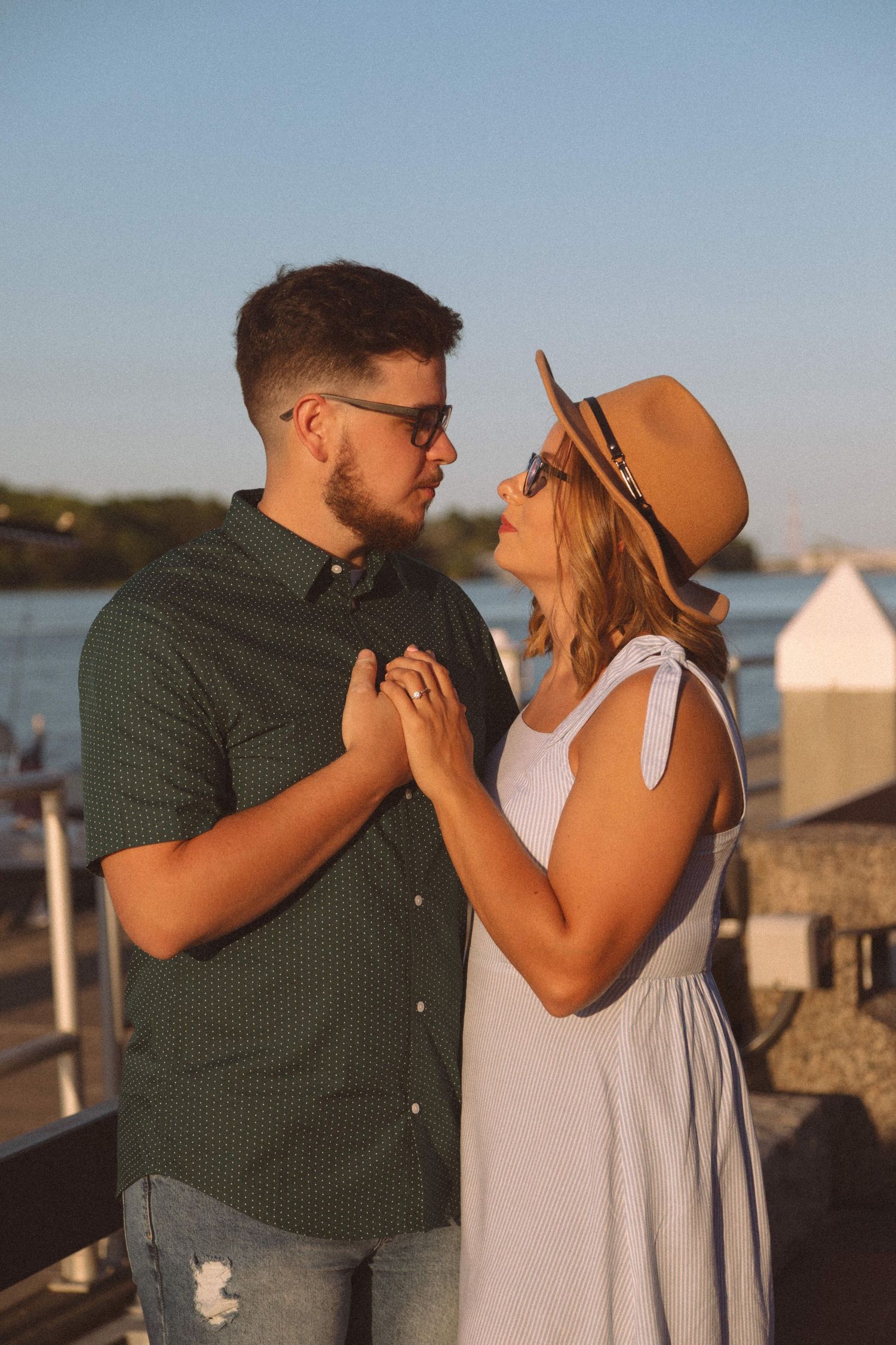 Couple holding hands and gazing into each other's eyes at sunset near the water, woman wearing hat and sunglasses.