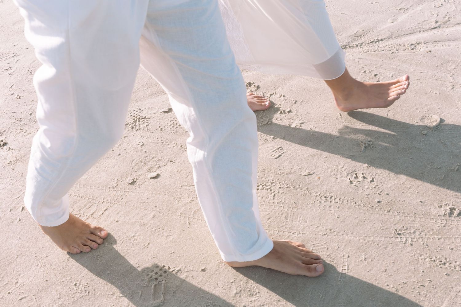 Barefoot person walking on white sand beach wearing white pants, close-up of feet and legs.