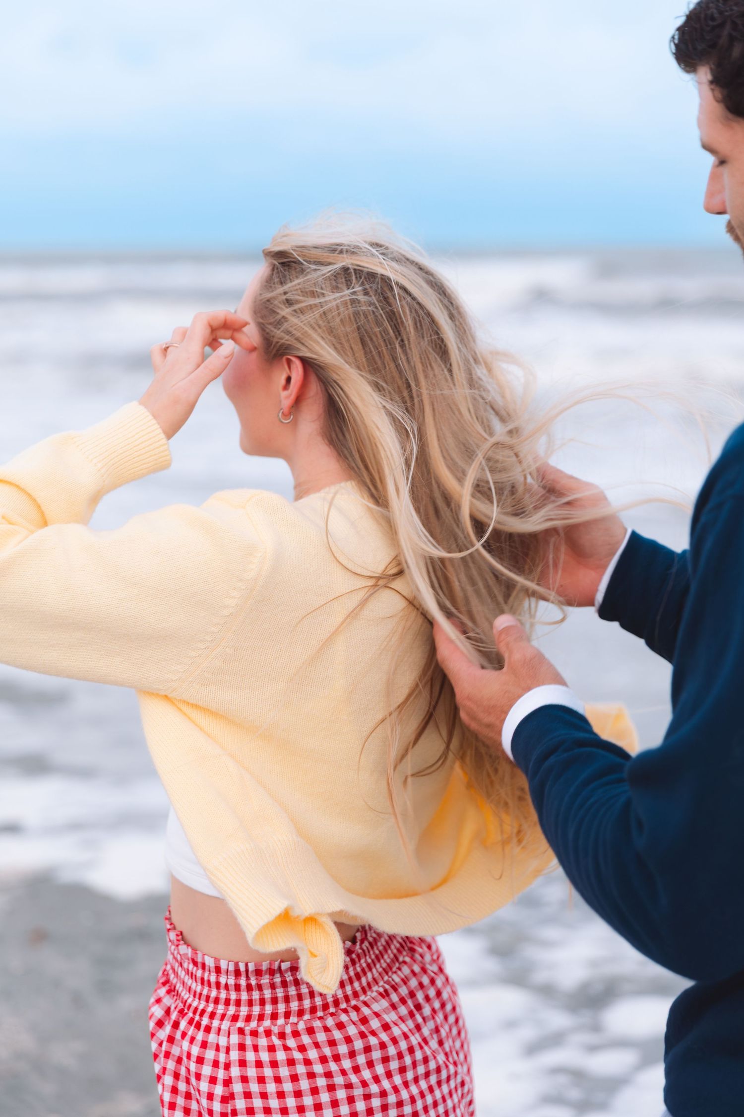 Man gently touching blonde womans windswept hair on a breezy beach with ocean waves.